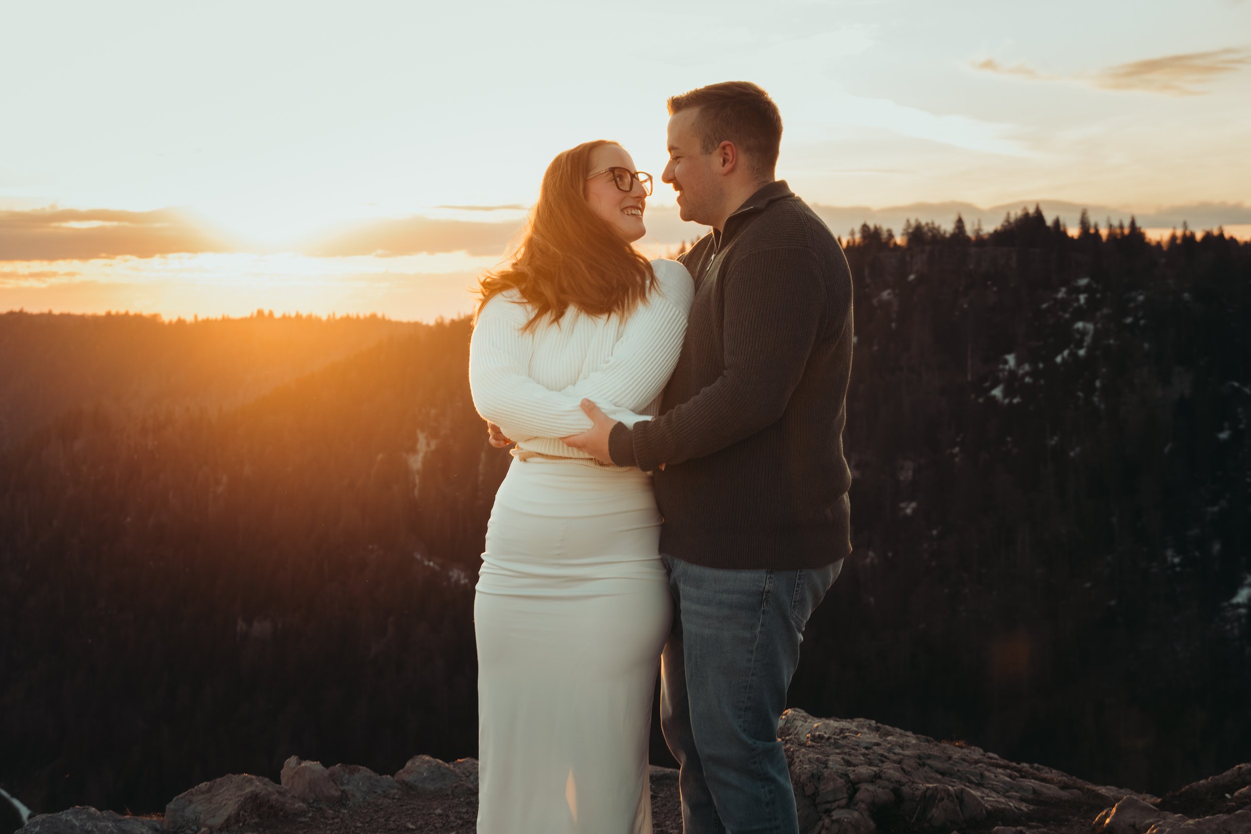 Un couple s'embrassant lors d'un coucher de soleil sur une montagne, avec des arbres en arrière-plan et des rochers au premier plan.