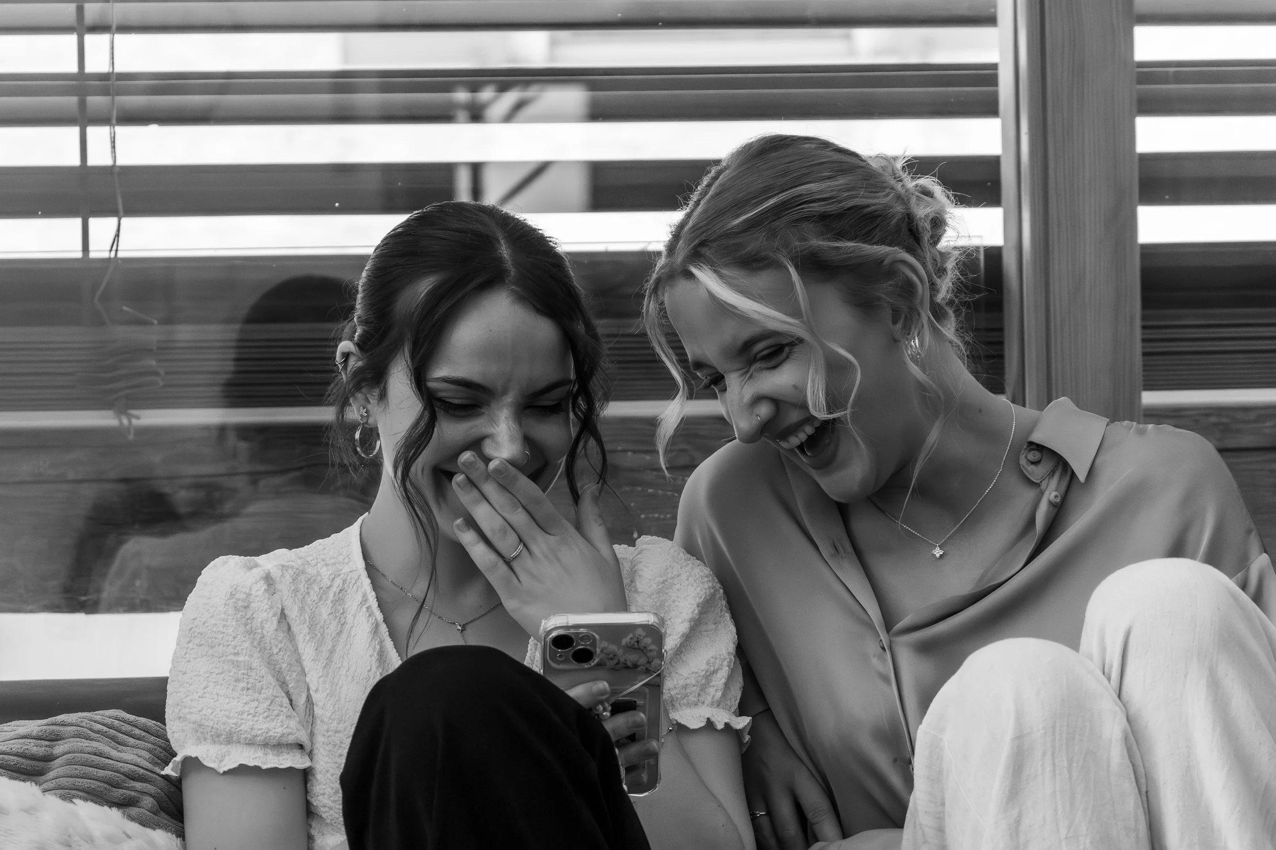 Deux femmes rient et prennent une photo avec un téléphone portable, assises côte à côte devant une structure en bois avec des volets horizontaux.