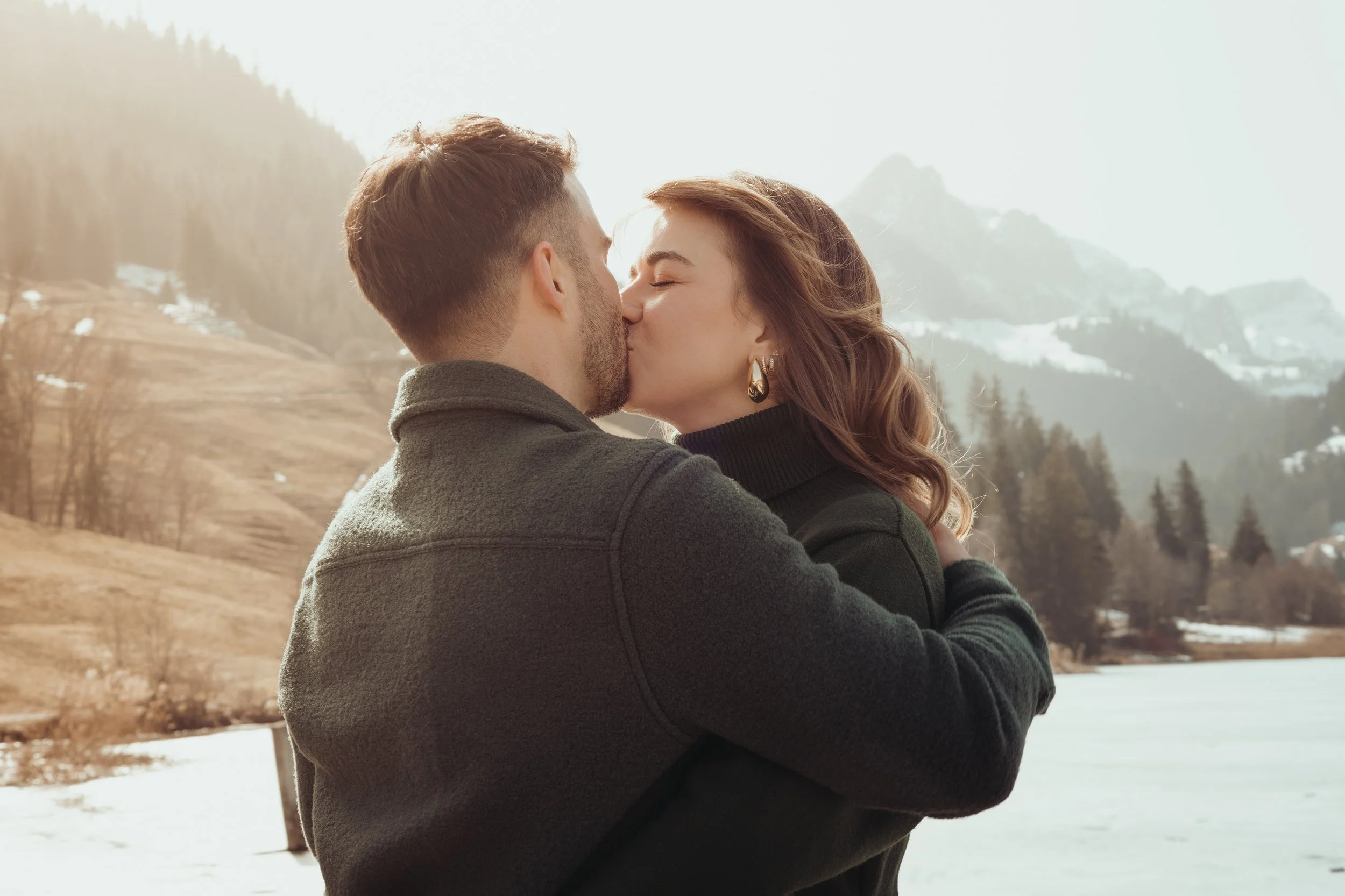 Un couple s'embrasse tendrement dans un paysage hivernal avec des montagnes et des arbres.