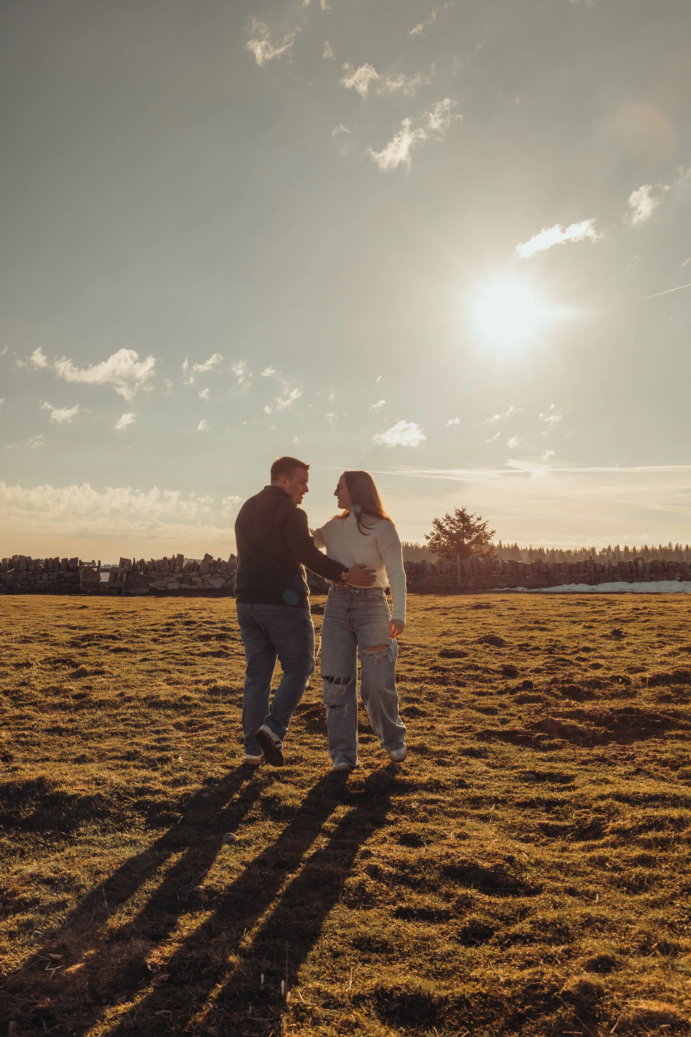 Un couple marche dans un champ en fin d'après-midi avec le soleil brillant dans le ciel, créant de longues ombres sur l'herbe.