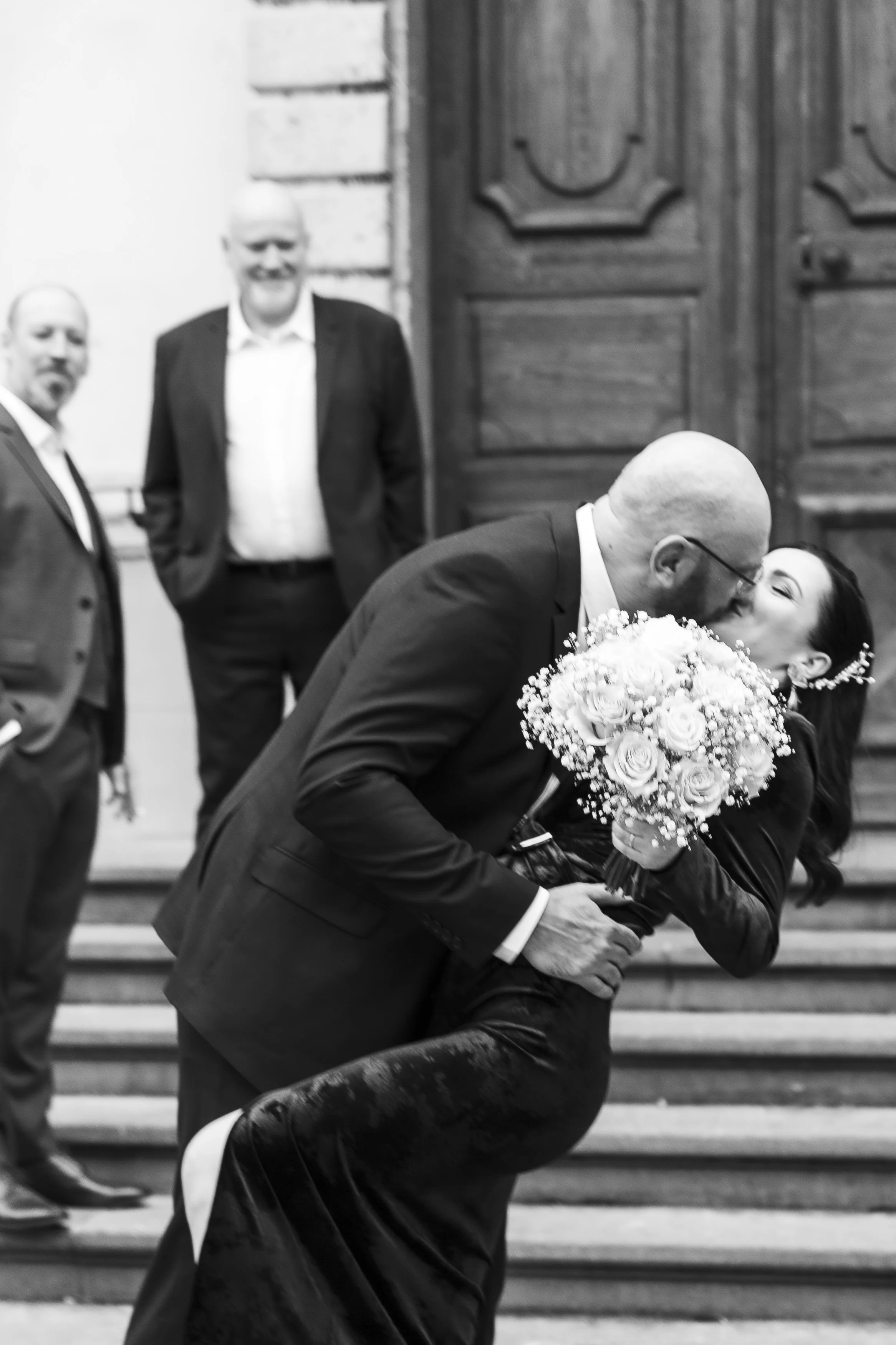 Un couple en mariage s'embrassant et s'enlaçant devant une porte en bois, entourés de témoins lors d'une cérémonie.