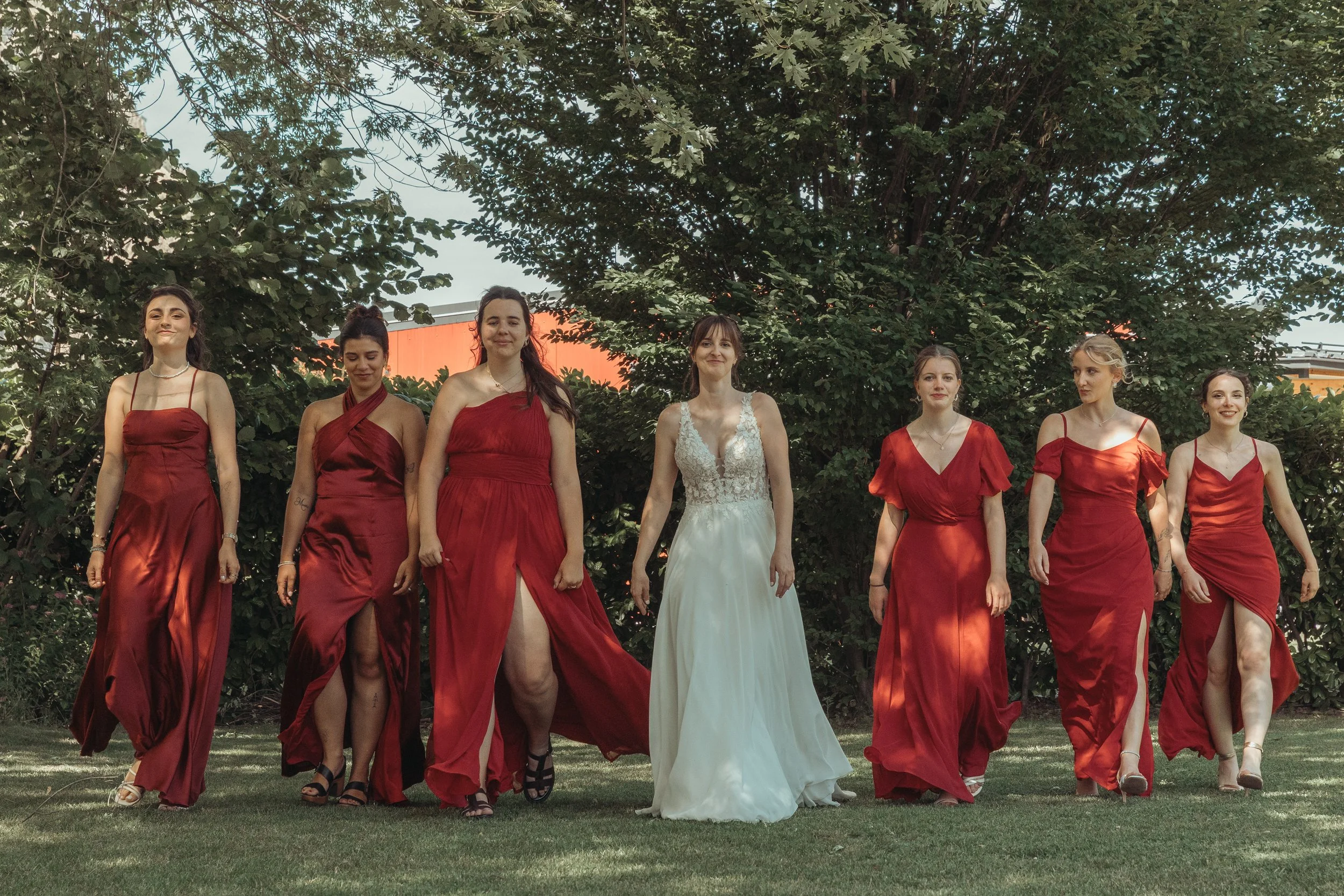 Un groupe de femmes habillées en tenues de soirée rouges et blanches, marchant ensemble dans un jardin avec des arbres en arrière-plan, lors d'une cérémonie ou d'une occasion spéciale.