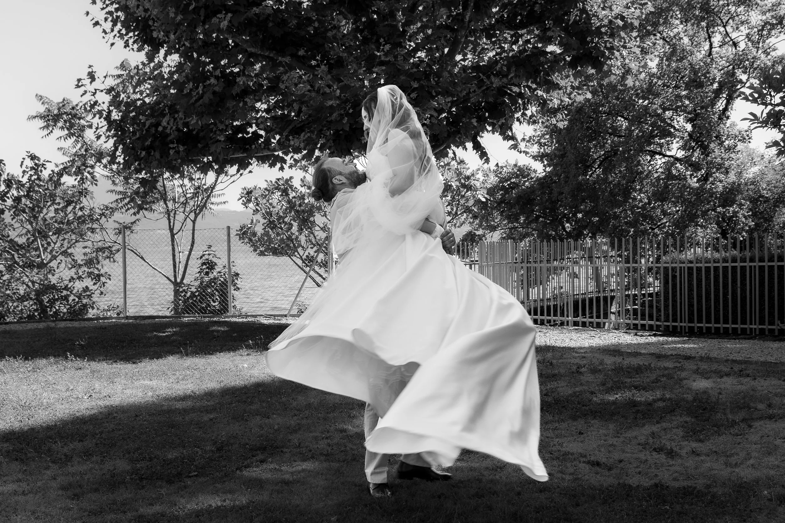 Un couple danse dans un parc, la femme en robe de mariage et le homme en costume, sous un arbre feuillu, en noir et blanc.
