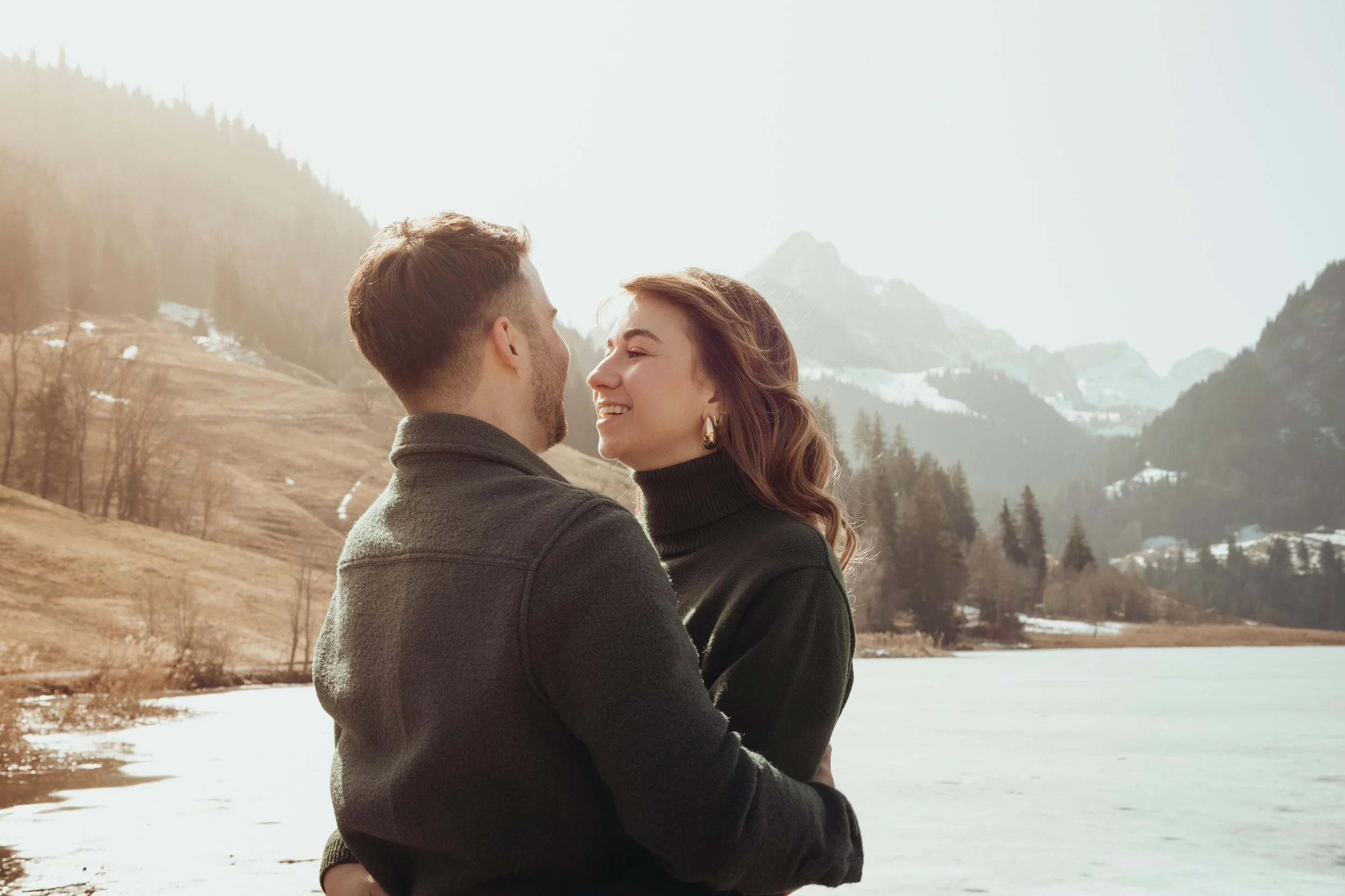 Un couple souriant face à face près d'un lac glacé en montagne, avec des collines et des arbres en arrière-plan, sous un ciel clair.