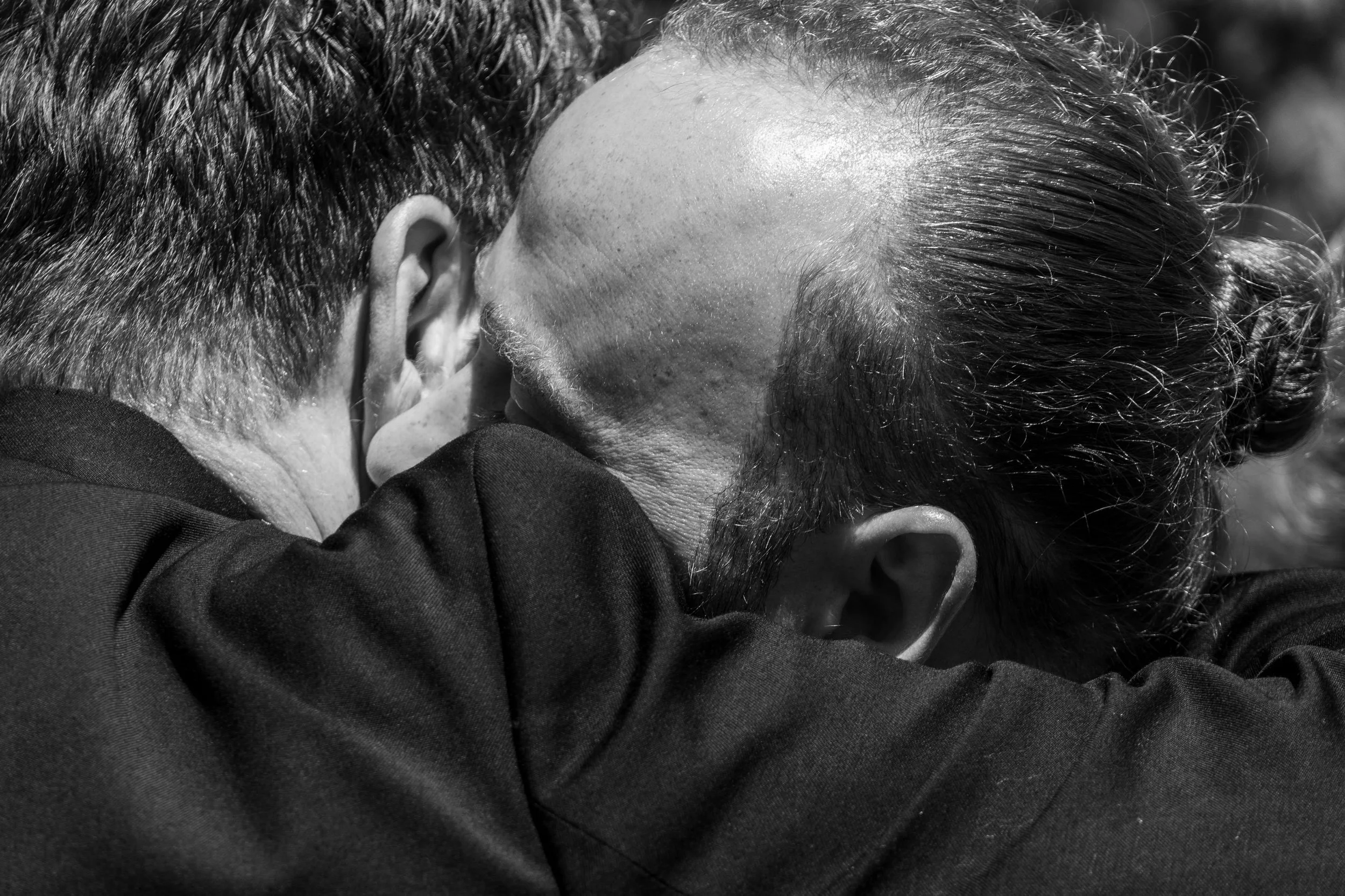 Deux personnes, visibles seulement de dos, s'embrassent étroitement, avec une personne ayant des cheveux courts et l'autre des cheveux longs, dans un environnement probablement extérieur.