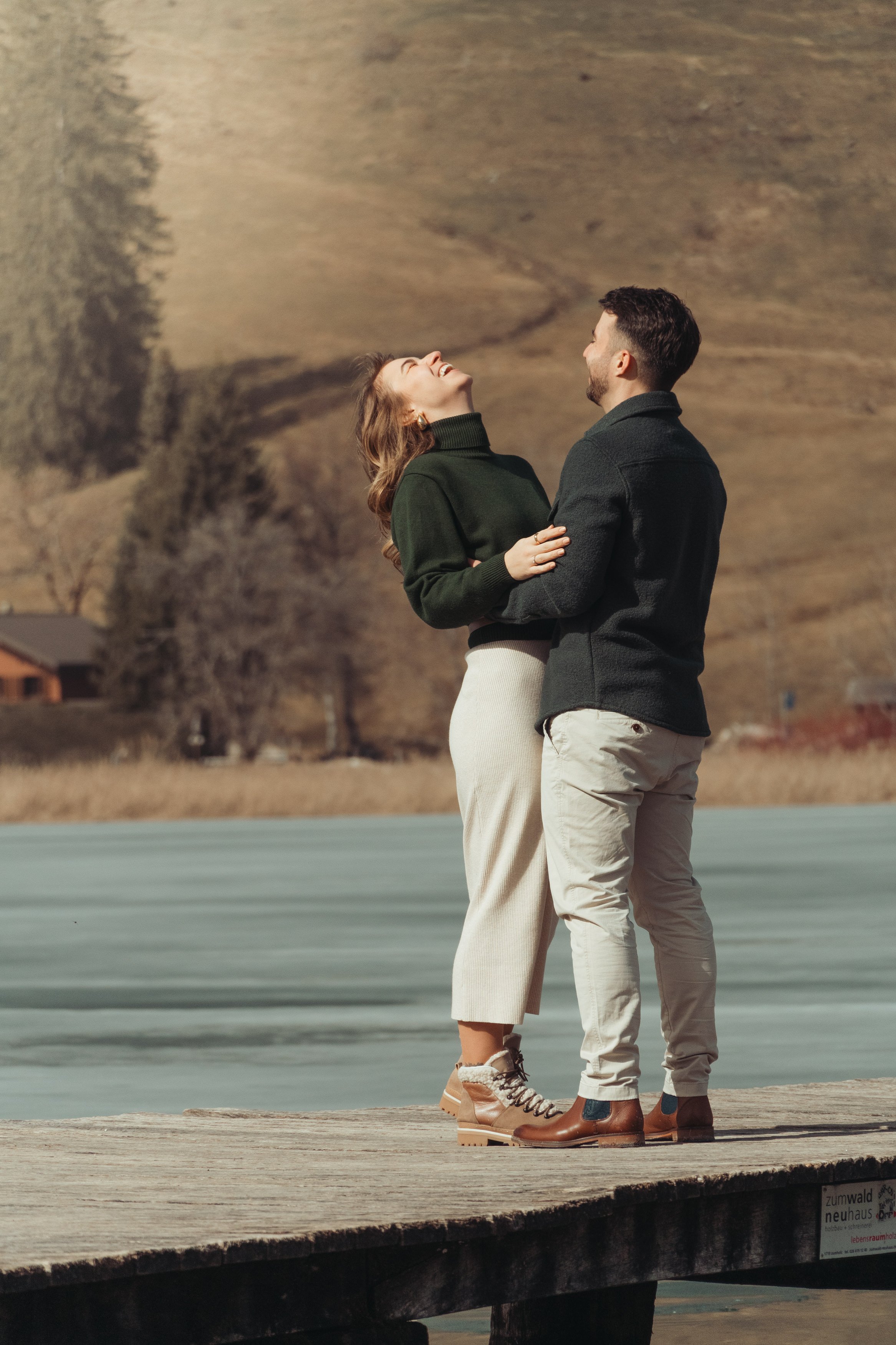 Un couple souriant se tenant par la taille, debout sur une jetée en bois, avec un fond de montagnes et de champs ouverts.
