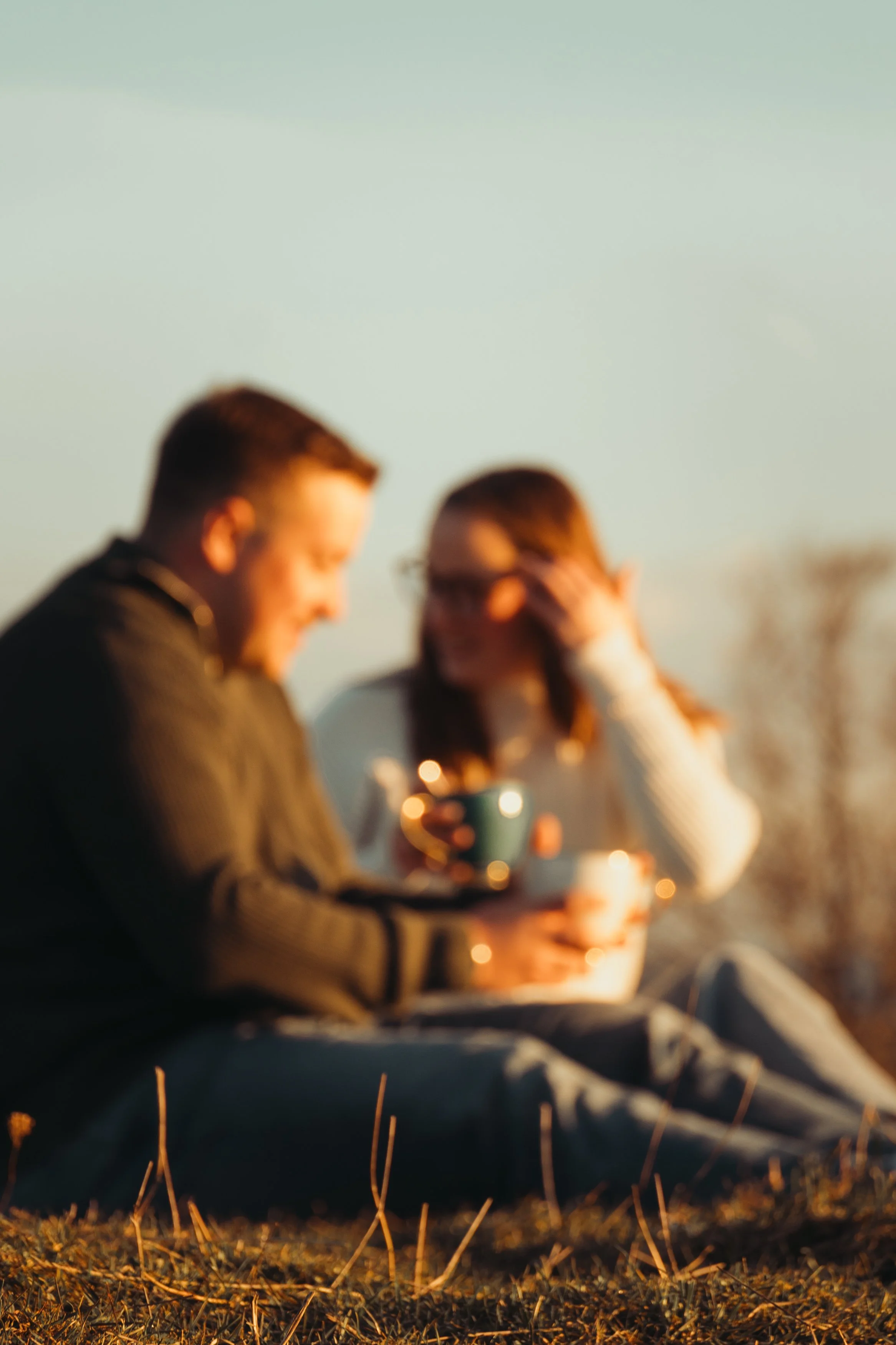 Deux personnes assises sur l'herbe lors d'un coucher de soleil, tenant des tasses à café ou à thé, avec une atmosphère chaleureuse et détendue.