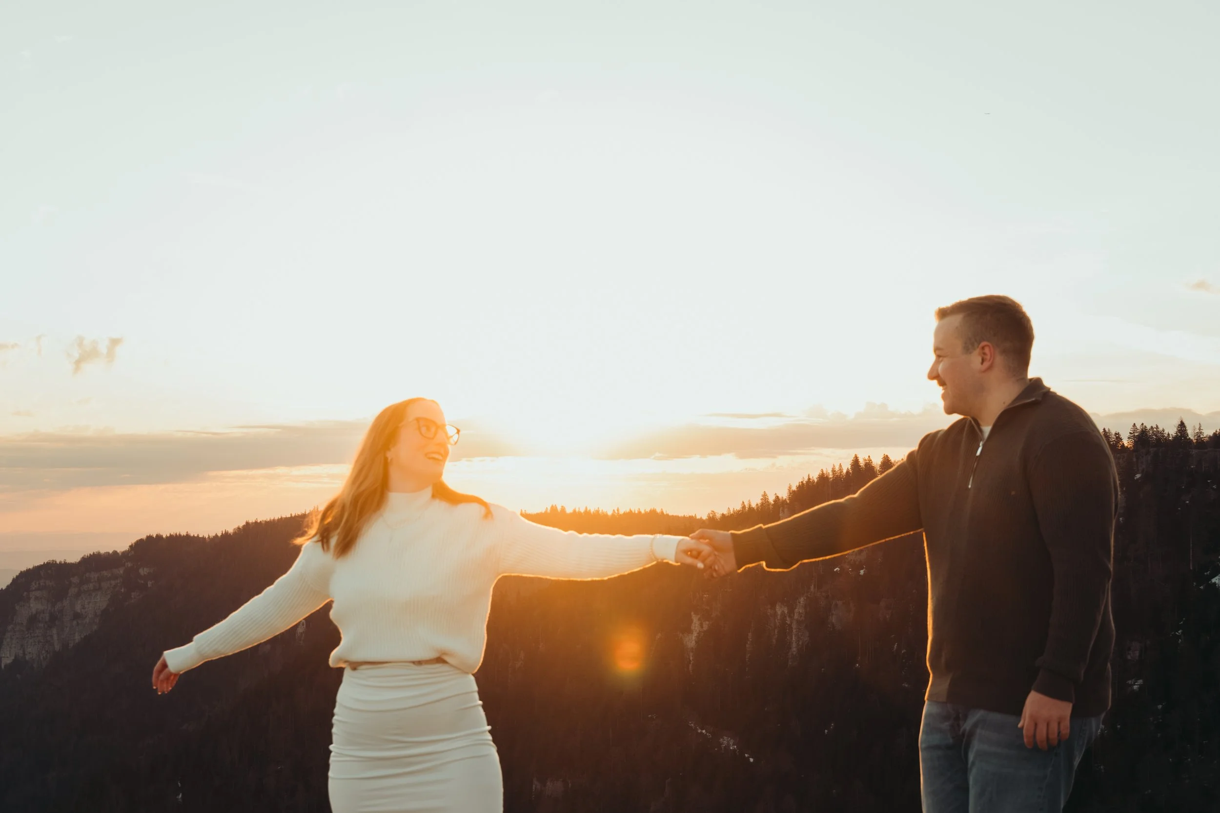 Un couple souriant se tient la main lors d'un coucher de soleil en plein air, dans un paysage de montagnes forestières.