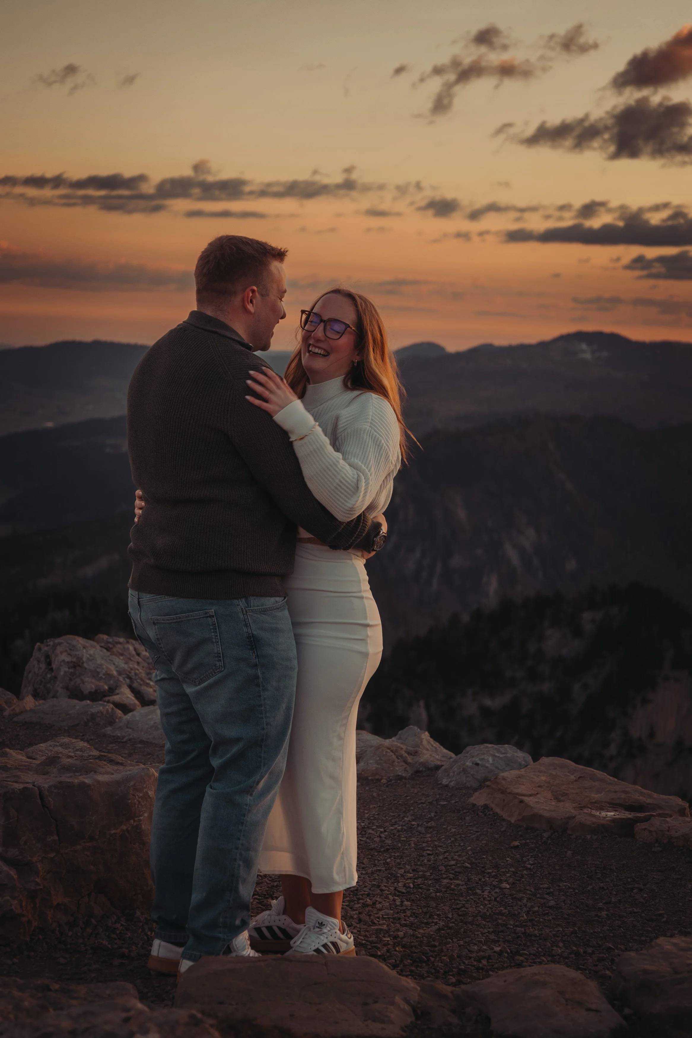 Un couple souriant et s'embrassant sur une montagne au coucher du soleil, avec un ciel orange et des nuages, des collines en arrière-plan.