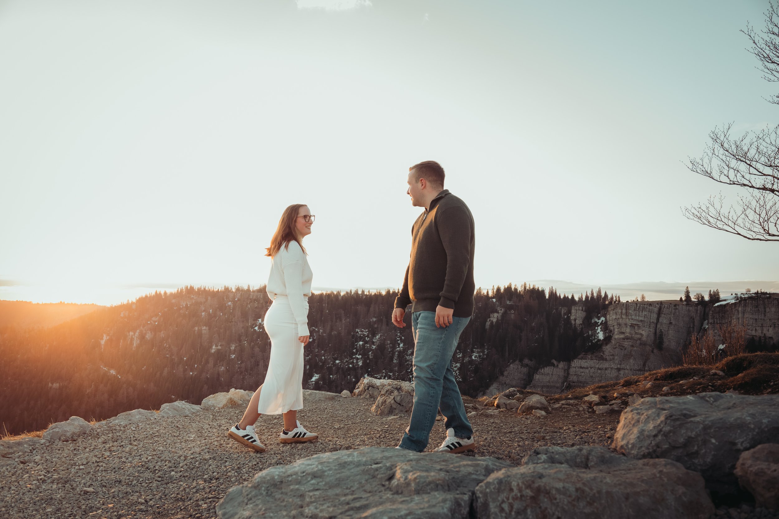 Un couple se tenant face à face en plein air au coucher du soleil, avec des montagnes en arrière-plan, des arbres nus de chaque côté, et des rochers au premier plan.