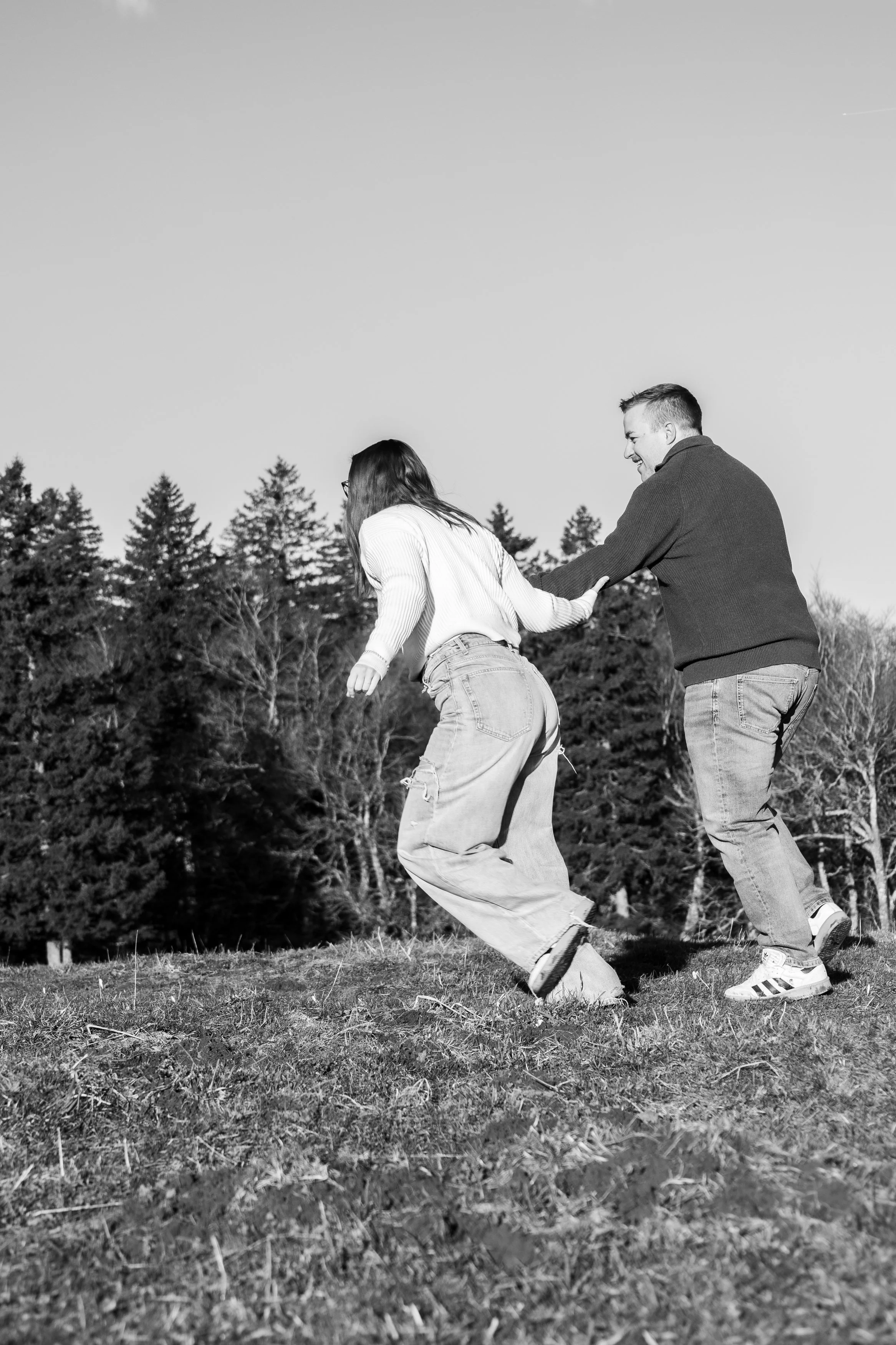 Deux personnes, un homme et une femme, jouent ensemble dans un parc ou une forêt, souriants et en mouvement, avec des arbres en arrière-plan. Photo en noir et blanc.