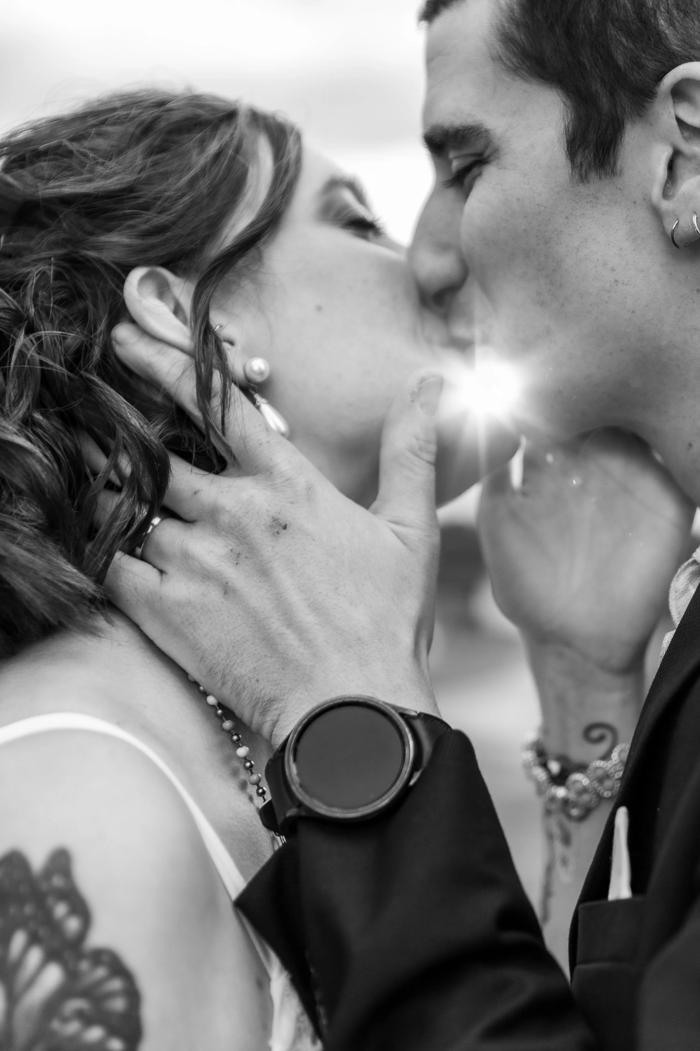 Deux personnes s'embrassent très près, avec leurs visages presque collés. L'image est en noir et blanc, la femme a des cheveux longs et bouclés, et porte des bijoux, la personne masculine porte des boucles d'oreilles et une montre smartwatch.