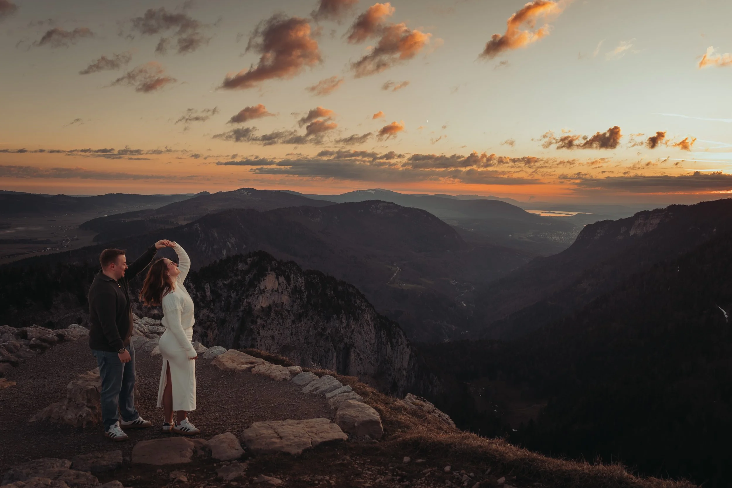 Un couple avec un guide au sommet d'une montagne au coucher du soleil, vue panoramique sur des montagnes et un lac à l'horizon, ciel avec des nuages orange et rose.