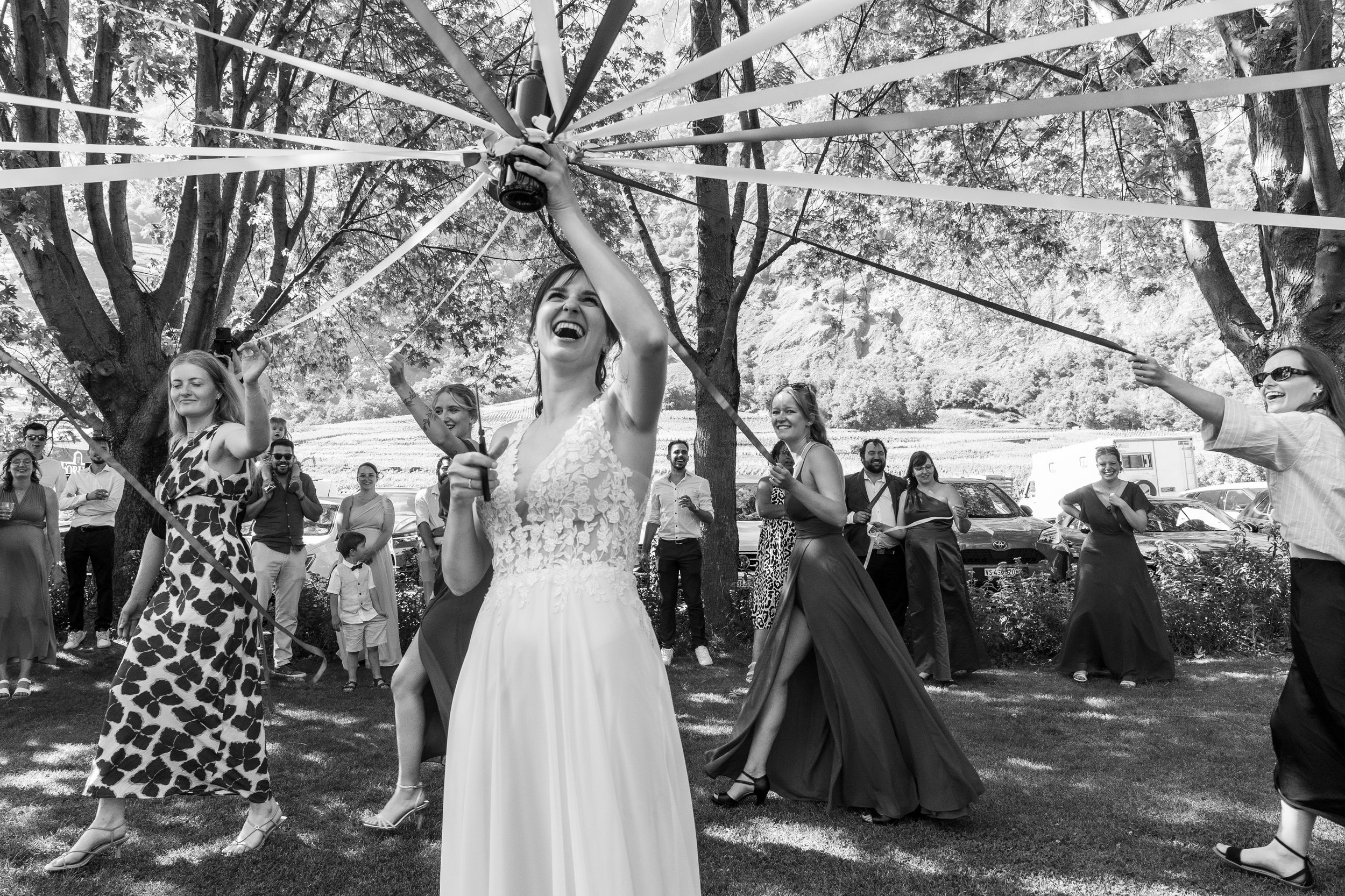 Un groupe de personnes danse et s'amuse lors d'une fête en plein air sous un arbre, avec une femme en robe blanche au centre, entourée d'autres femmes en robes de différentes couleurs et de spectateurs souriants.