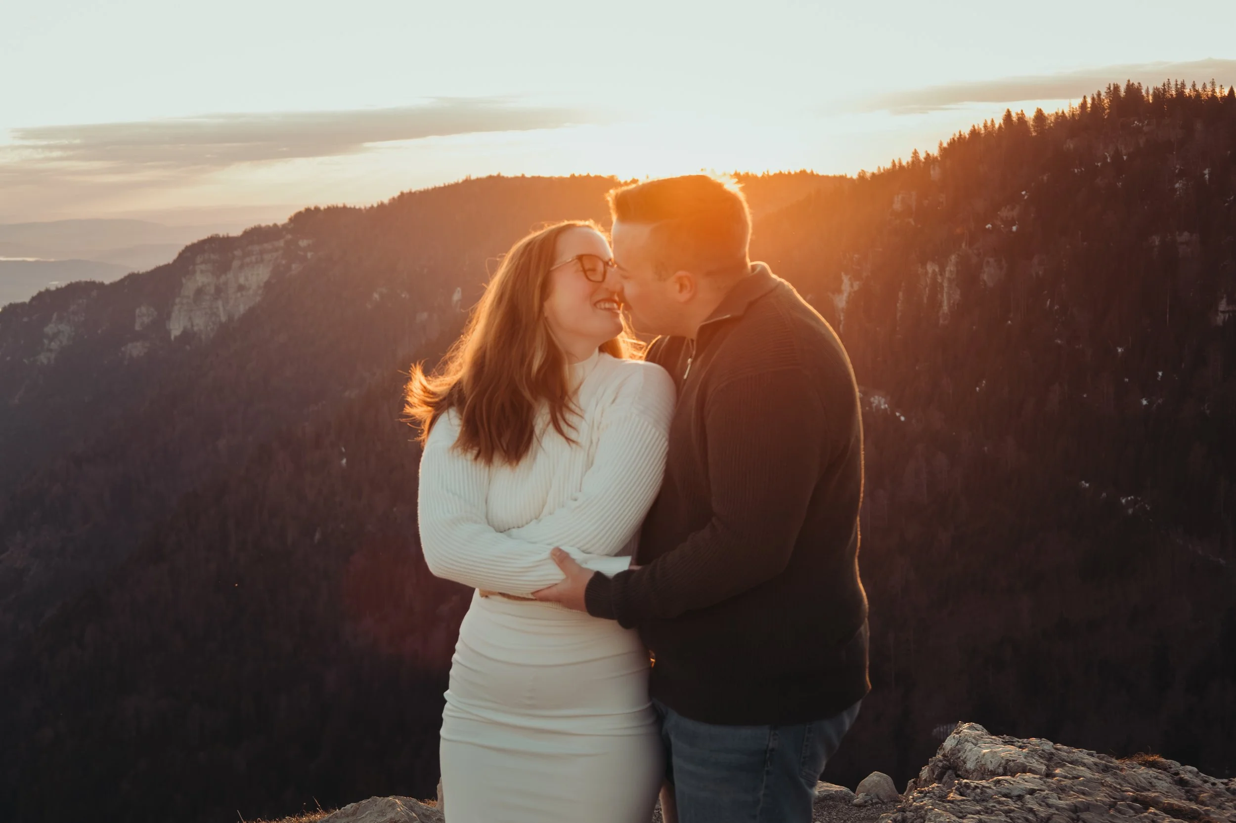 Un couple souriant, en embrassant, avec un paysage de montagnes au coucher du soleil en arrière-plan.
