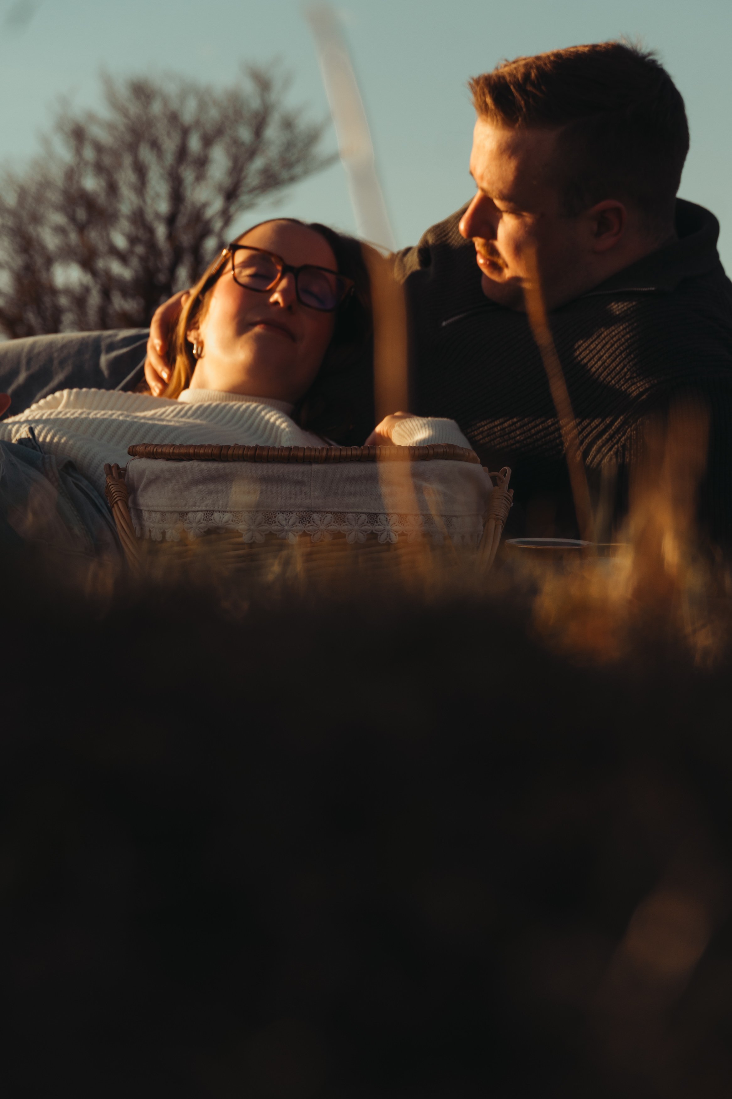 Un couple assis près d'un feu de camp lors du coucher du soleil, la femme porte des lunettes et sourit, l'homme regarde vers elle. En arrière-plan, un arbre sans feuilles et un ciel lumineux sont visibles.