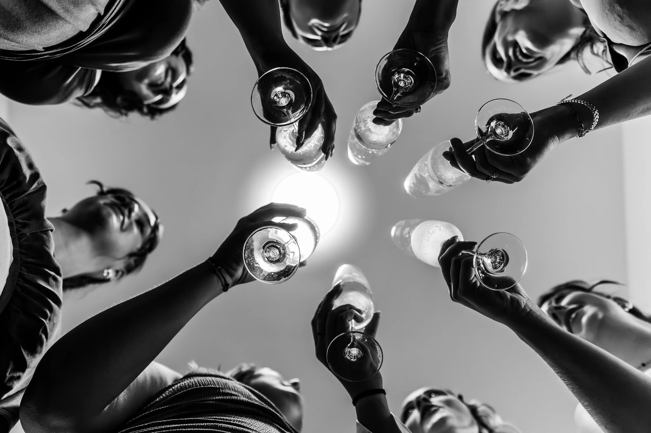 Groupe de personnes levant des verres à vin lors d'une célébration, vue de dessous en noir et blanc.