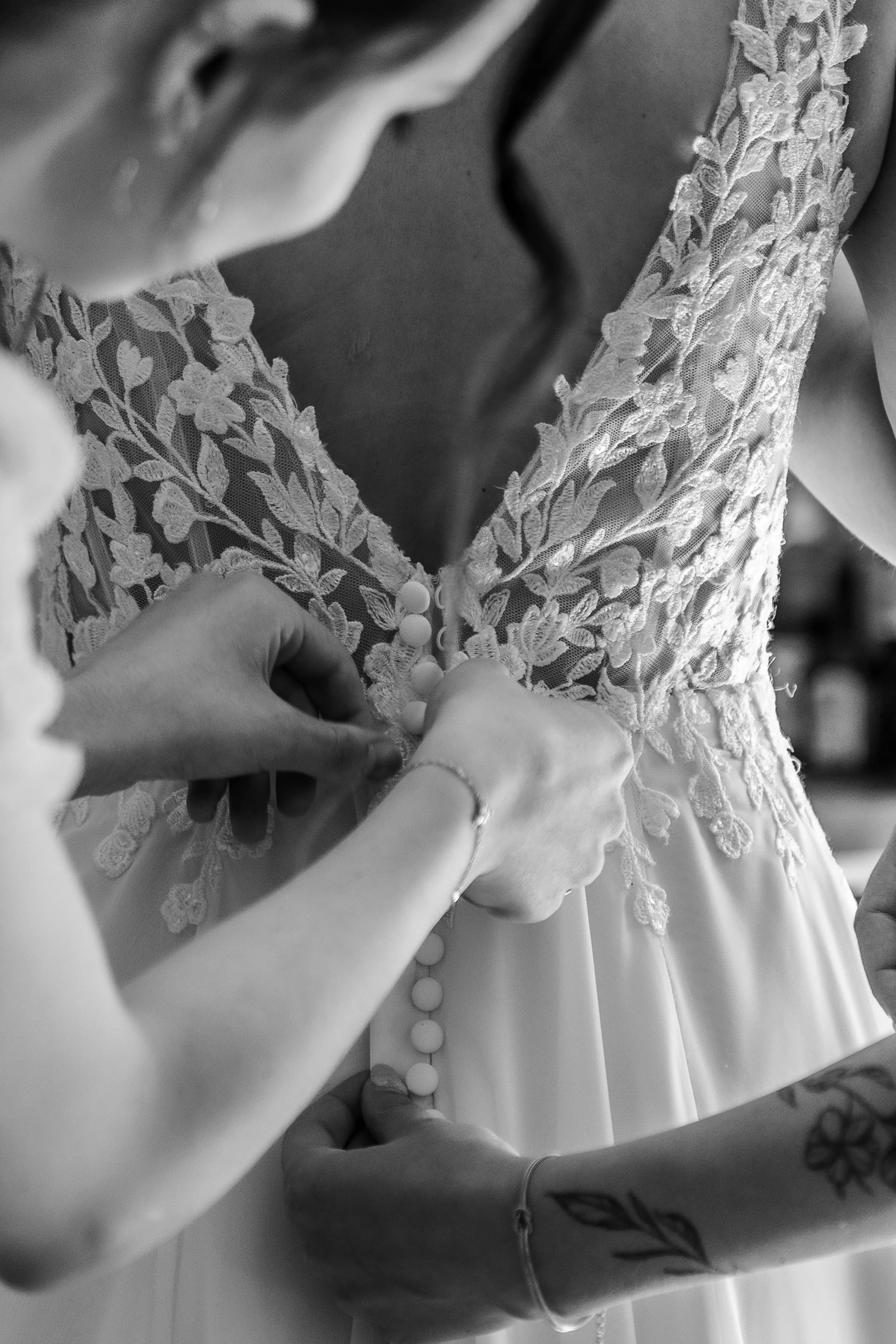 Détail d'une robe de mariée avec des broderies florales, des perles et une personne en train de boutonner la robe, avec d'autres mains aidant.