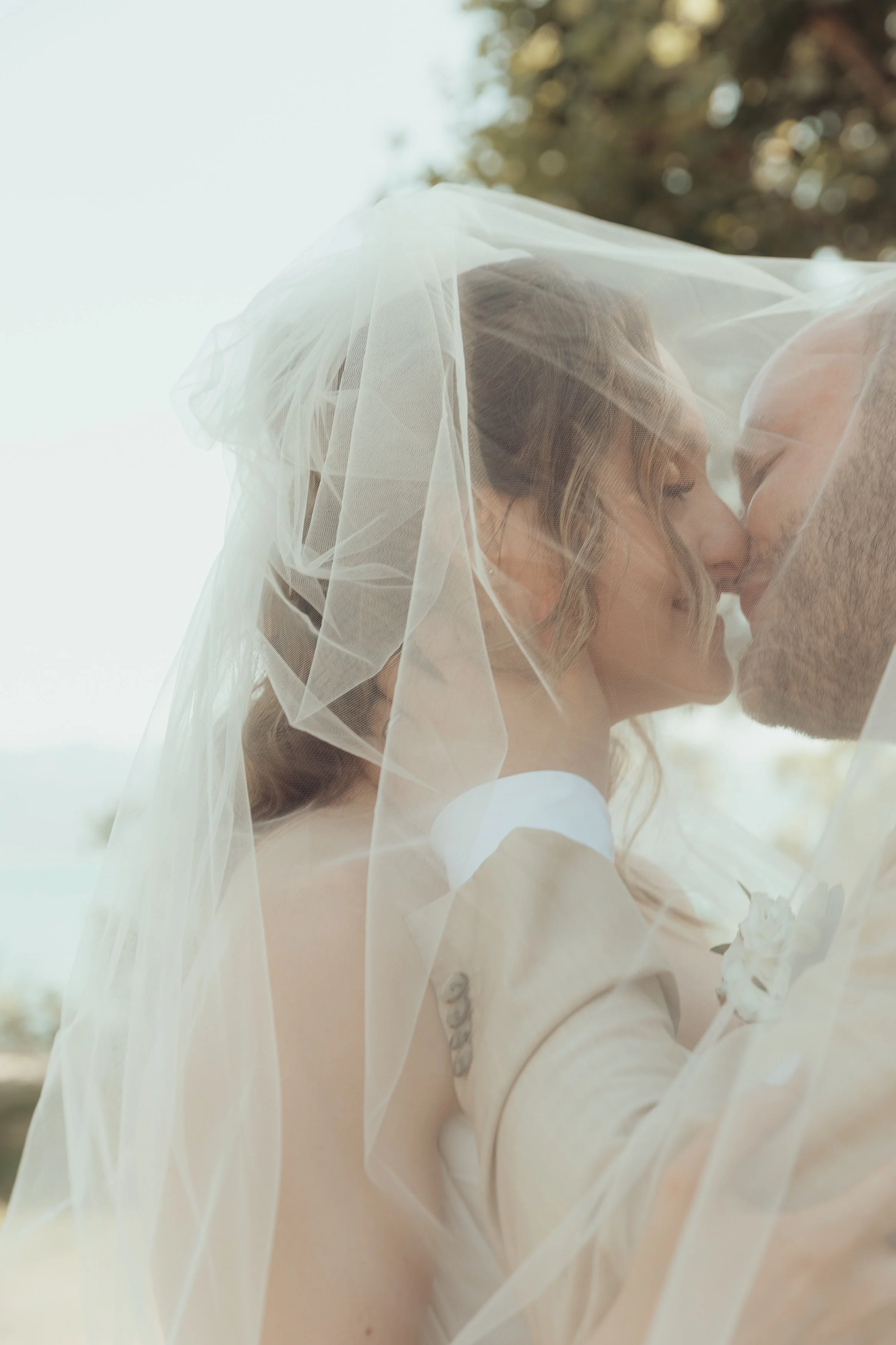 Un couple s'embrasse tendrement, vêtus de vêtements de mariage, avec une mariée portant un voile transparent au-dessus d'eux lors d'une séance photo en extérieur.