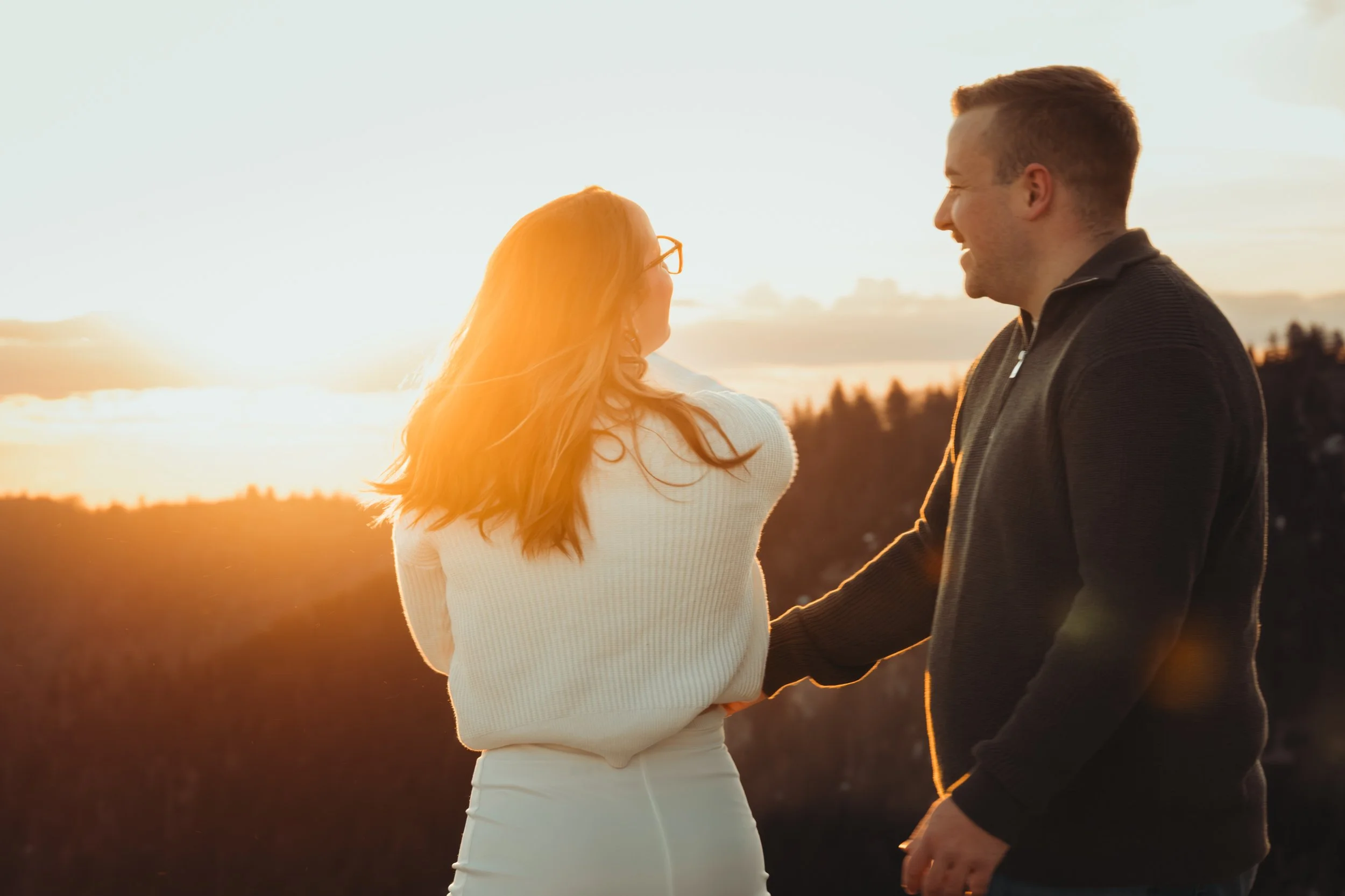 Un couple jeune, homme et femme, se tiennent la main et se regardent avec le sourire lors d'un coucher de soleil à l'extérieur.