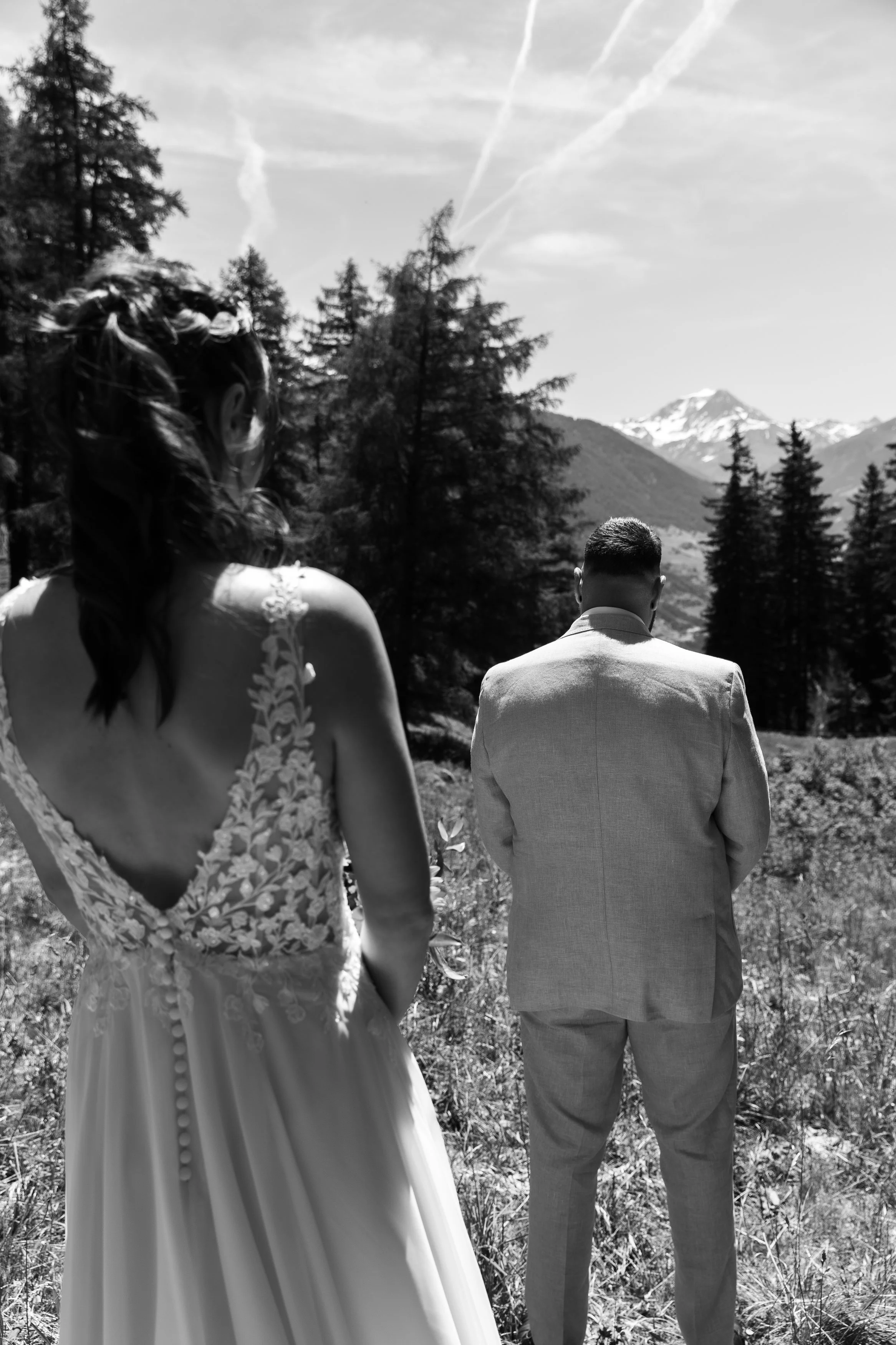 Deux personnes habillées pour un mariage en montagne, avec des montagnes enneigées en arrière-plan et des arbres autour, photographiées en noir et blanc.
