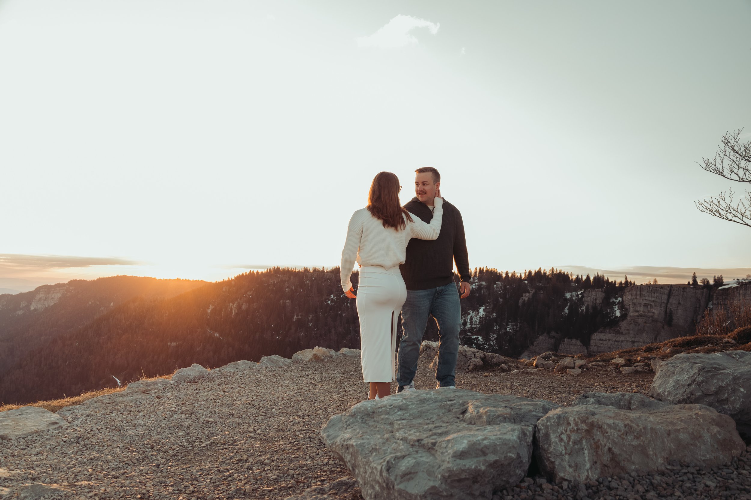 Un couple avec un homme et une femme se tenant par la main, se regardant, dans un paysage au coucher du soleil, avec des collines et des arbres dénudés en arrière-plan.