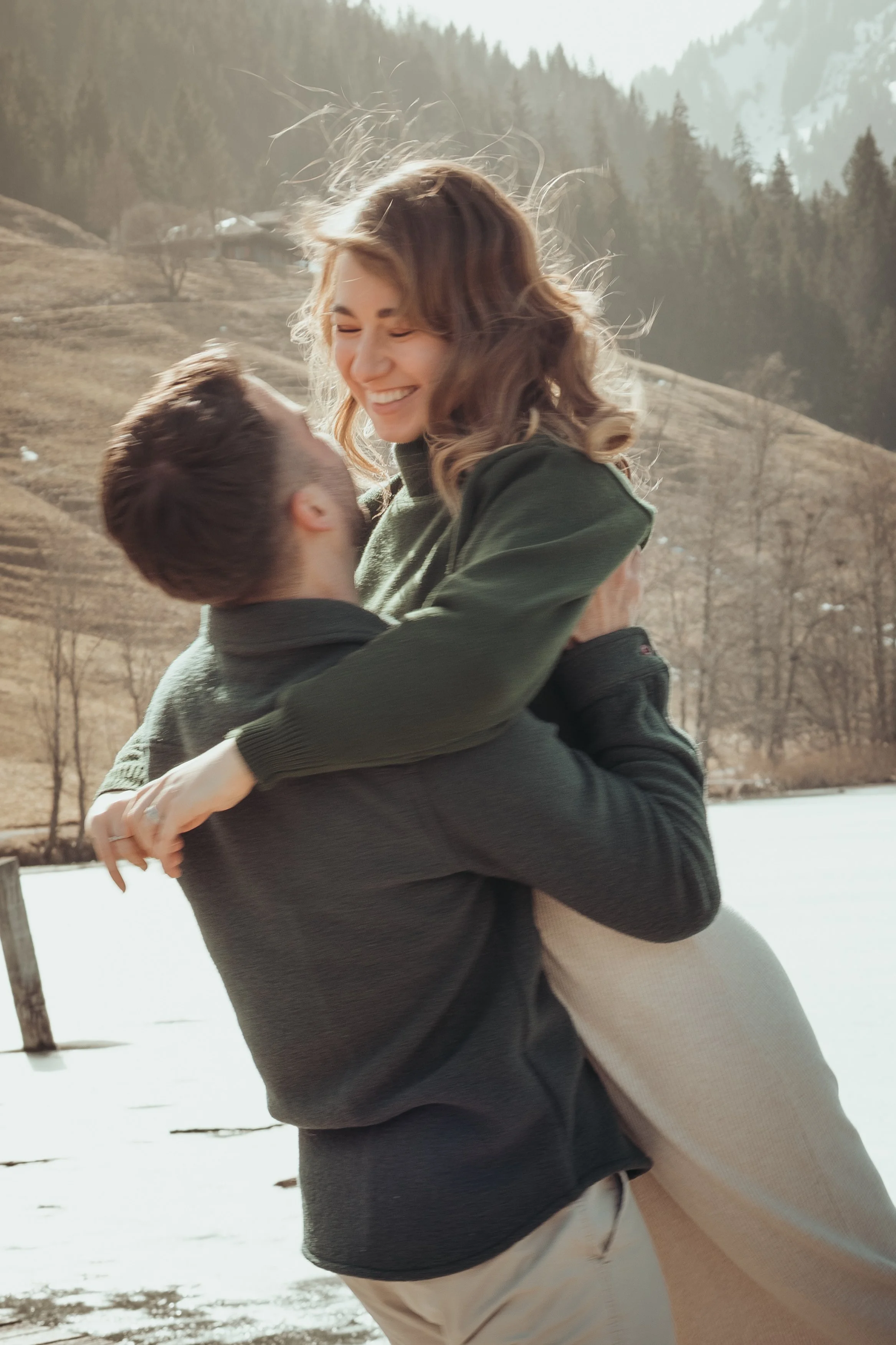 Une femme souriante portant un pull gris clair et un pantalon beige, étant levée dans les bras d'un homme. Ils sont en plein air près d'un lac ou d'une rivière, avec des montagnes et des arbres en arrière-plan.