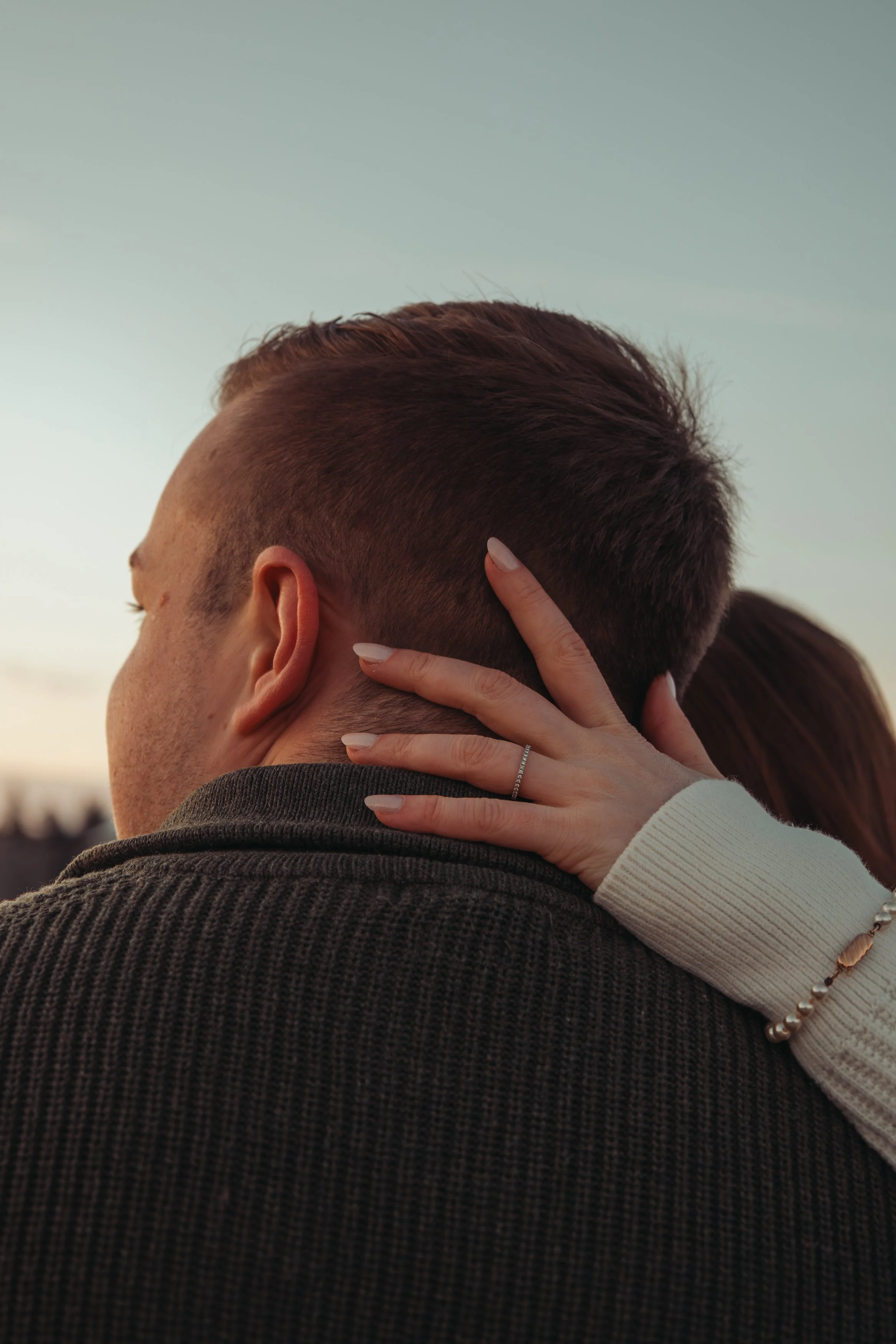 Un couple s'embrasse ou s'étreint, avec la personne derrière ayant une coiffure courte et la main sur la nuque de l'autre. La scène est prise lors d'un coucher de soleil ou dans une lumière douce.