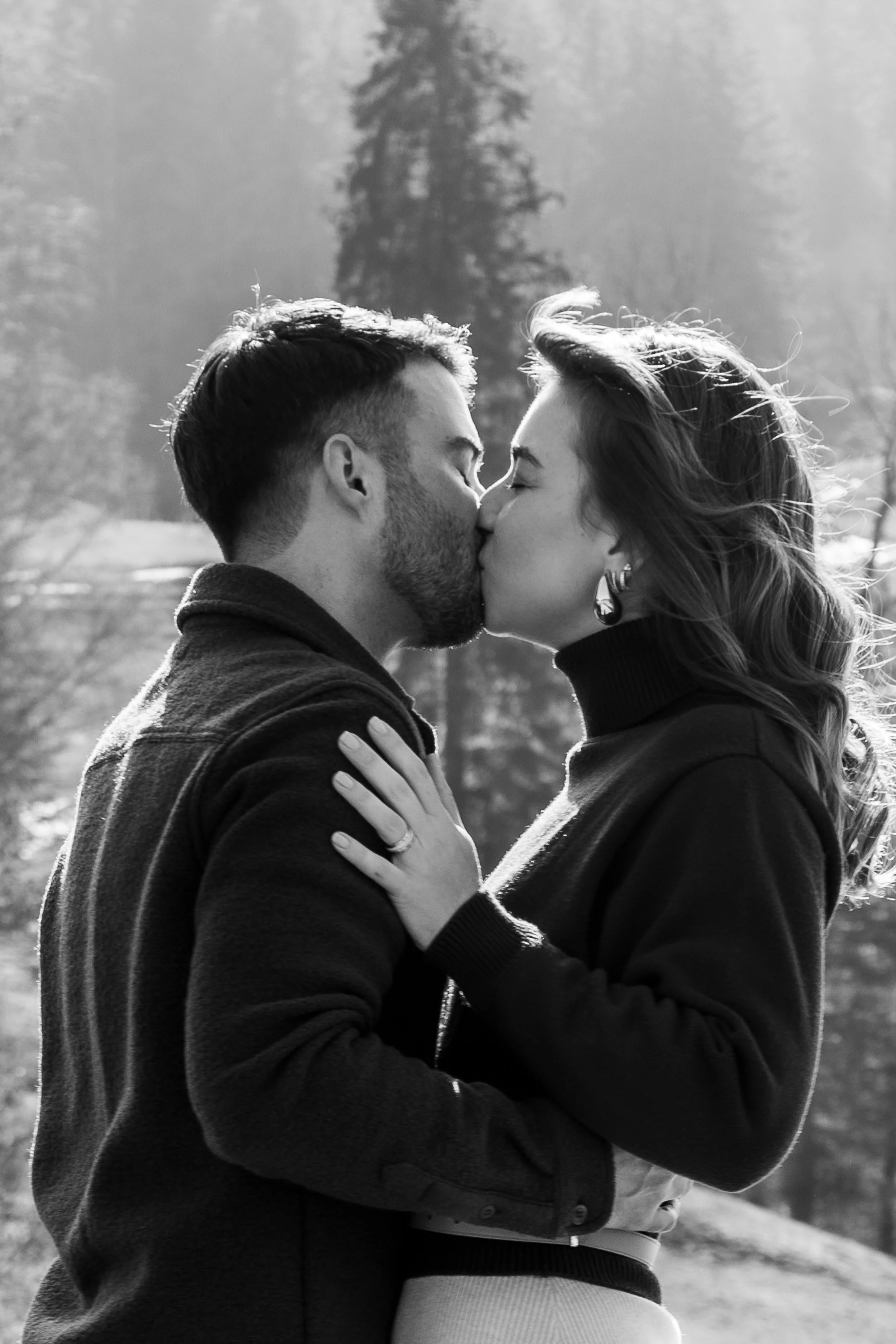 Deux personnes, un homme et une femme, s'embrassent dans un parc en plein air avec des arbres en arrière-plan.