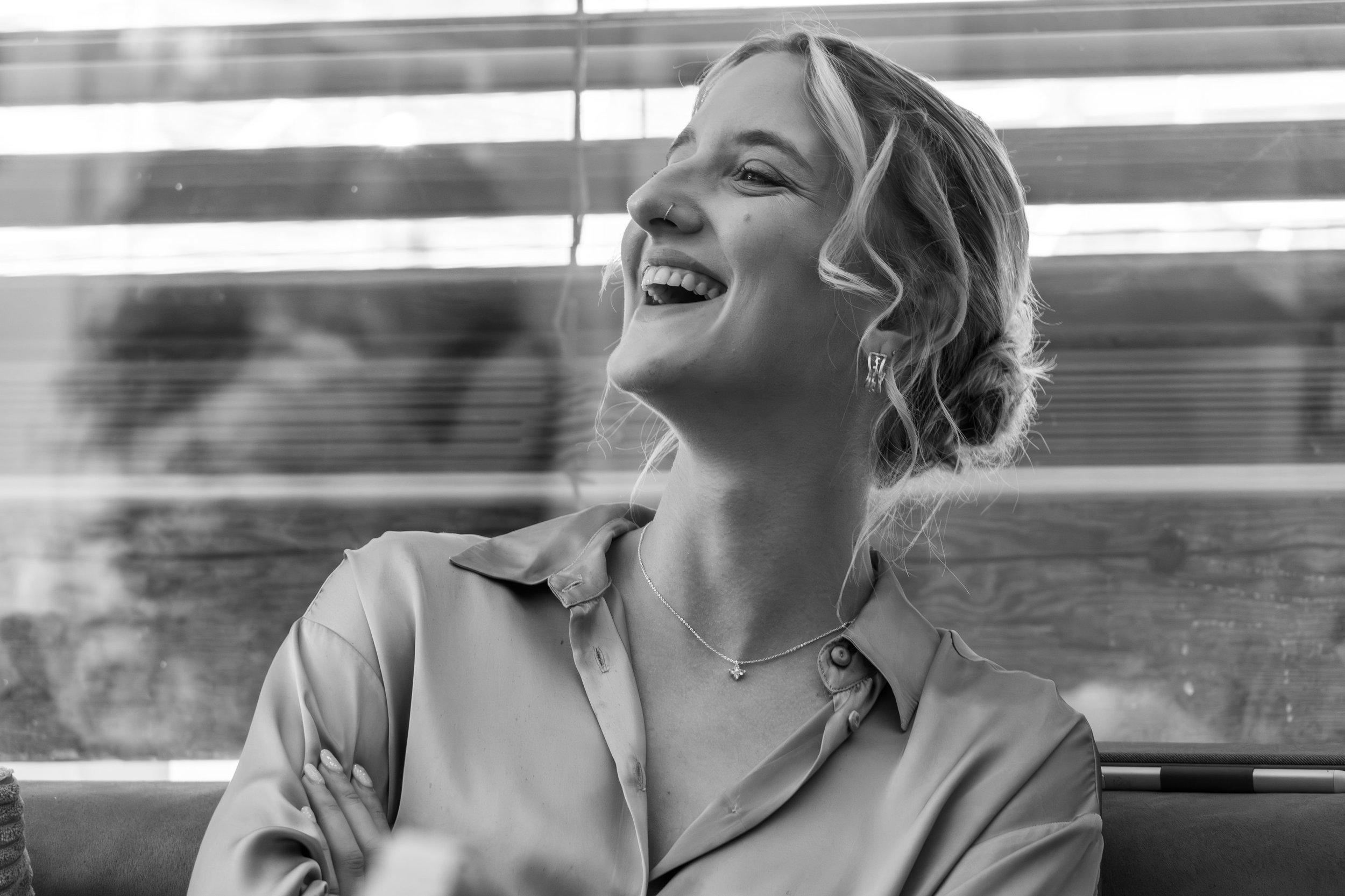 Jeune femme souriante et détendue, portant un collier, assise sur un banc en bois, plissant légèrement les yeux, dans un environnement lumineux avec des volets ou un bardage en bois en arrière-plan.