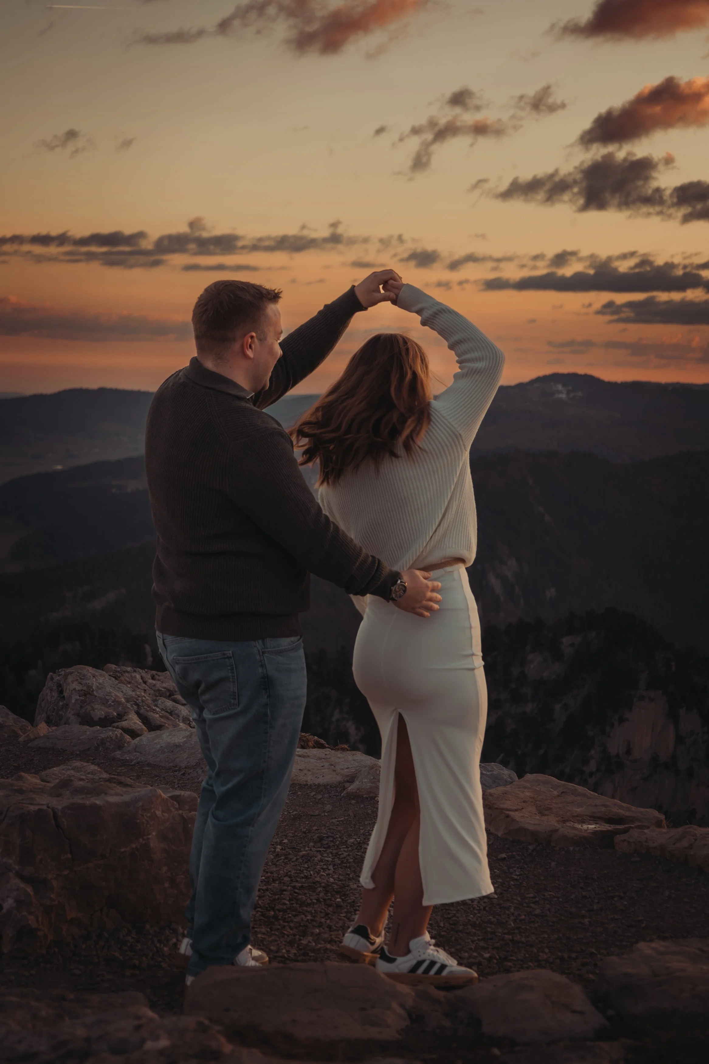 Un couple danse sur un rocher au coucher du soleil, avec un paysage montagneux en arrière-plan.