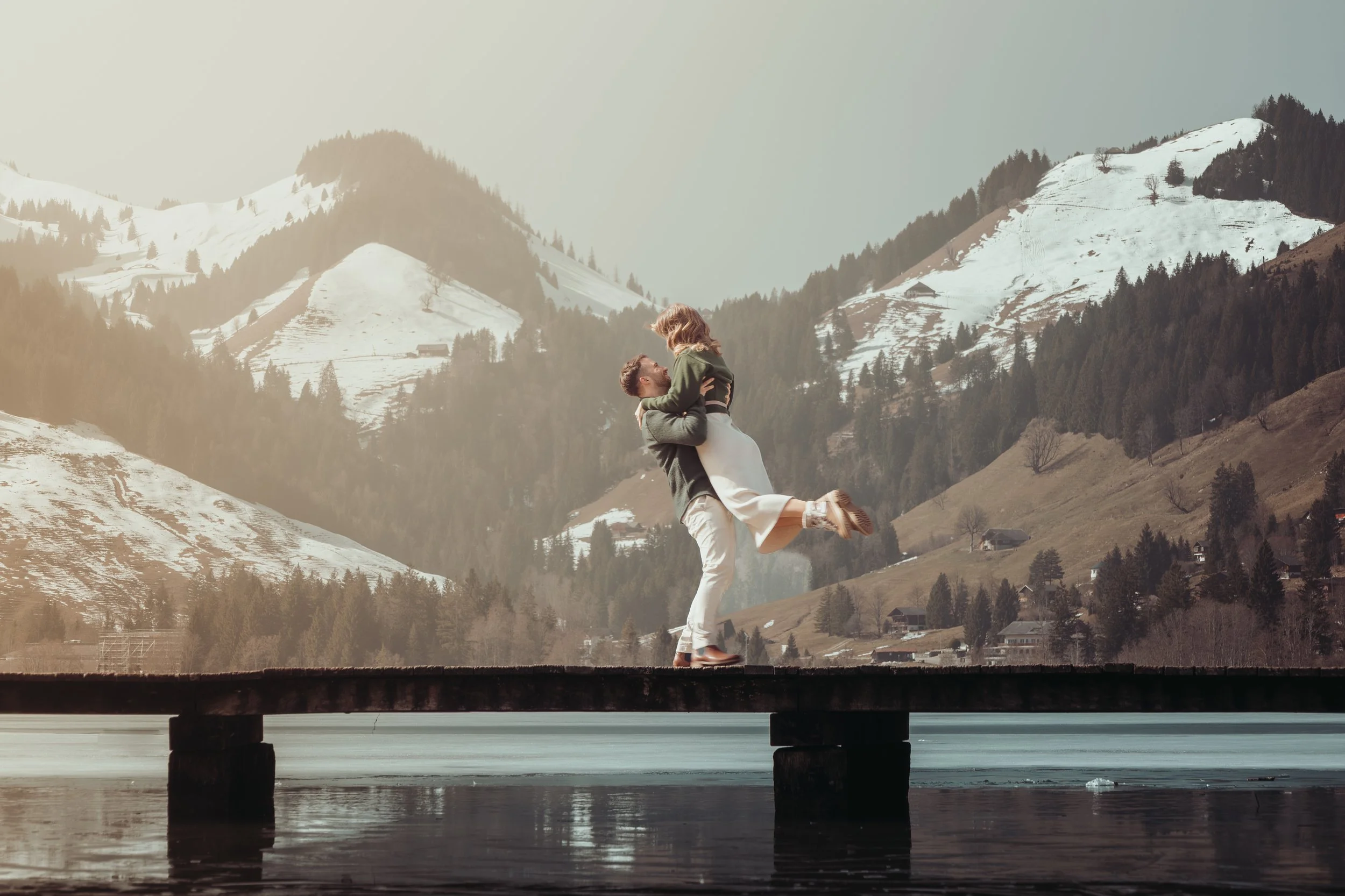 Un couple danse joyeusement sous un pont en bois au bord d'un lac, avec des montagnes enneigées en arrière-plan, dans un paysage pittoresque.