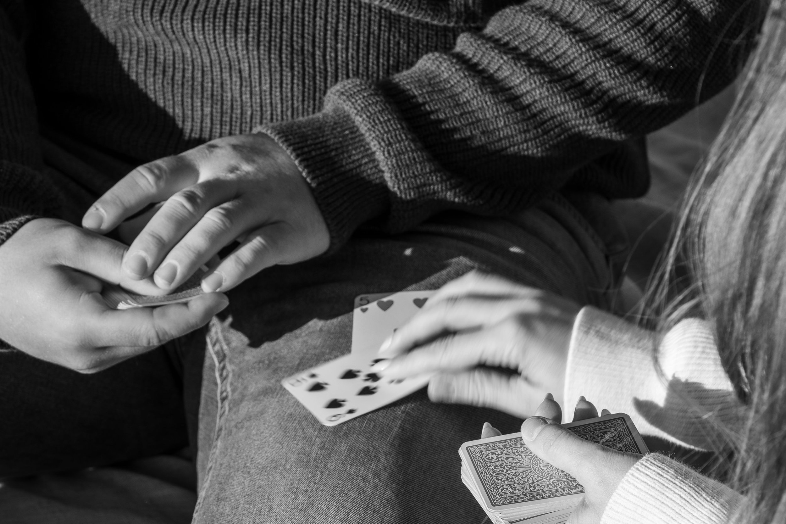 Deux personnes jouent aux cartes, l'une d'elles tient un paquet de cartes françaises en main, une autre a une main avec une carte visible, toutes deux portent des pulls en laine. L'image est en noir et blanc.