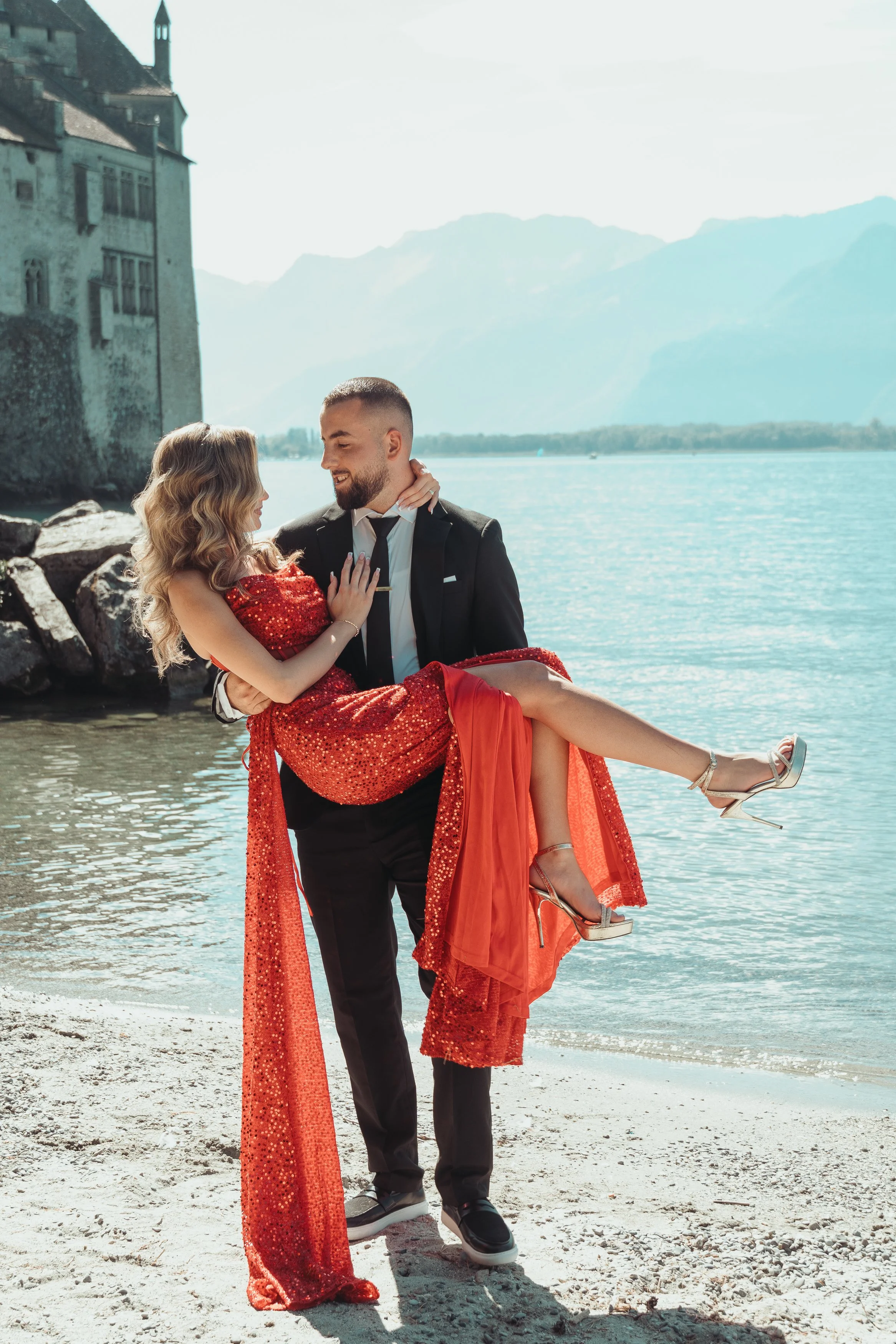 Un homme en costume noir tient une femme en robe rouge pailletée, près d'un lac avec un château en arrière-plan, dans un paysage de montagnes lors d'une séance photo couple en Suisse avec Marie Monod photographe d'émotions, duo de photographes queer