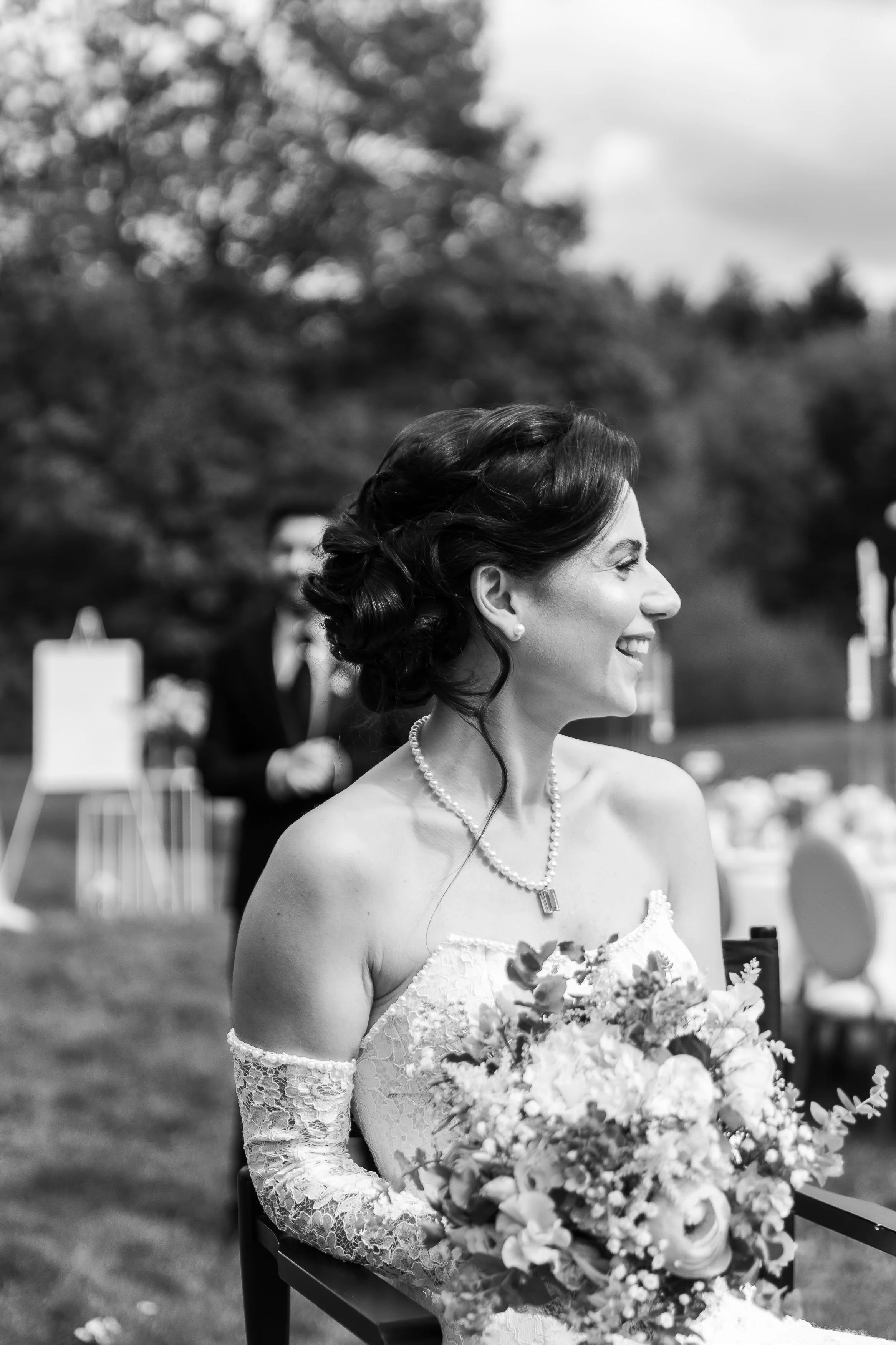 Portrait en noir et blanc d'une femme souriante vêtue d'une robe de mariage en dentelle, portant un collier de perles, tenant un bouquet de fleurs, à l'extérieur.