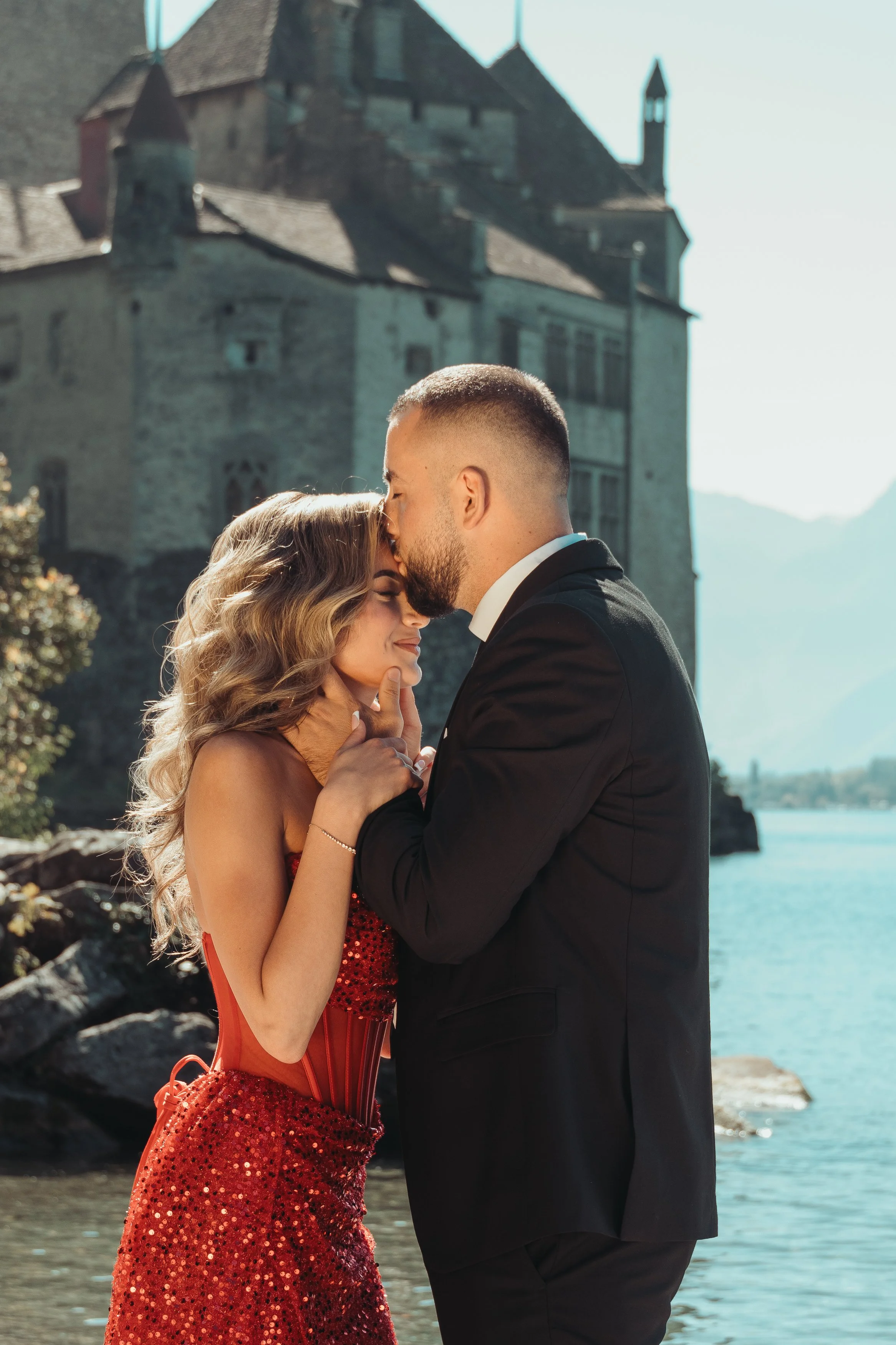 Un couple habillé élégamment, un homme en costume noir et une femme en robe rouge, partage un moment romantique près de l'eau avec un château en pierre en arrière-plan lors d'une séance photo avec Marie Monod photographe d'émotions