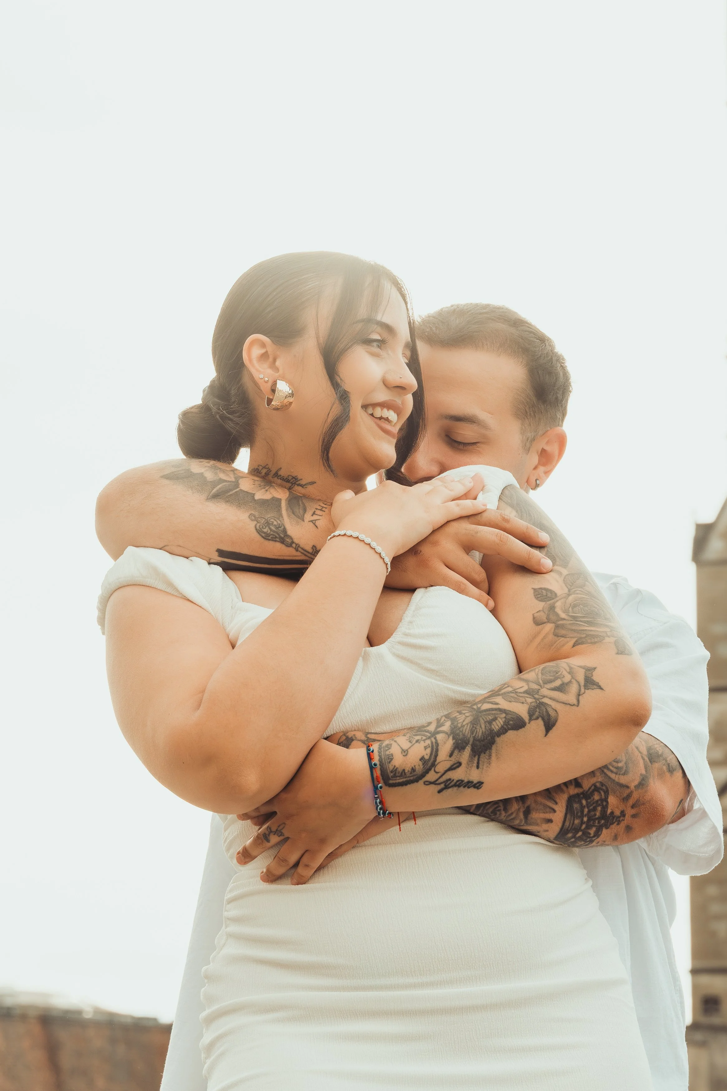 Un couple heureux s'embrassant et se blottant l'un contre l'autre en extérieur, avec un ciel clair en fond, la femme souriant avec des tatouages visibles sur ses bras et son épaule.