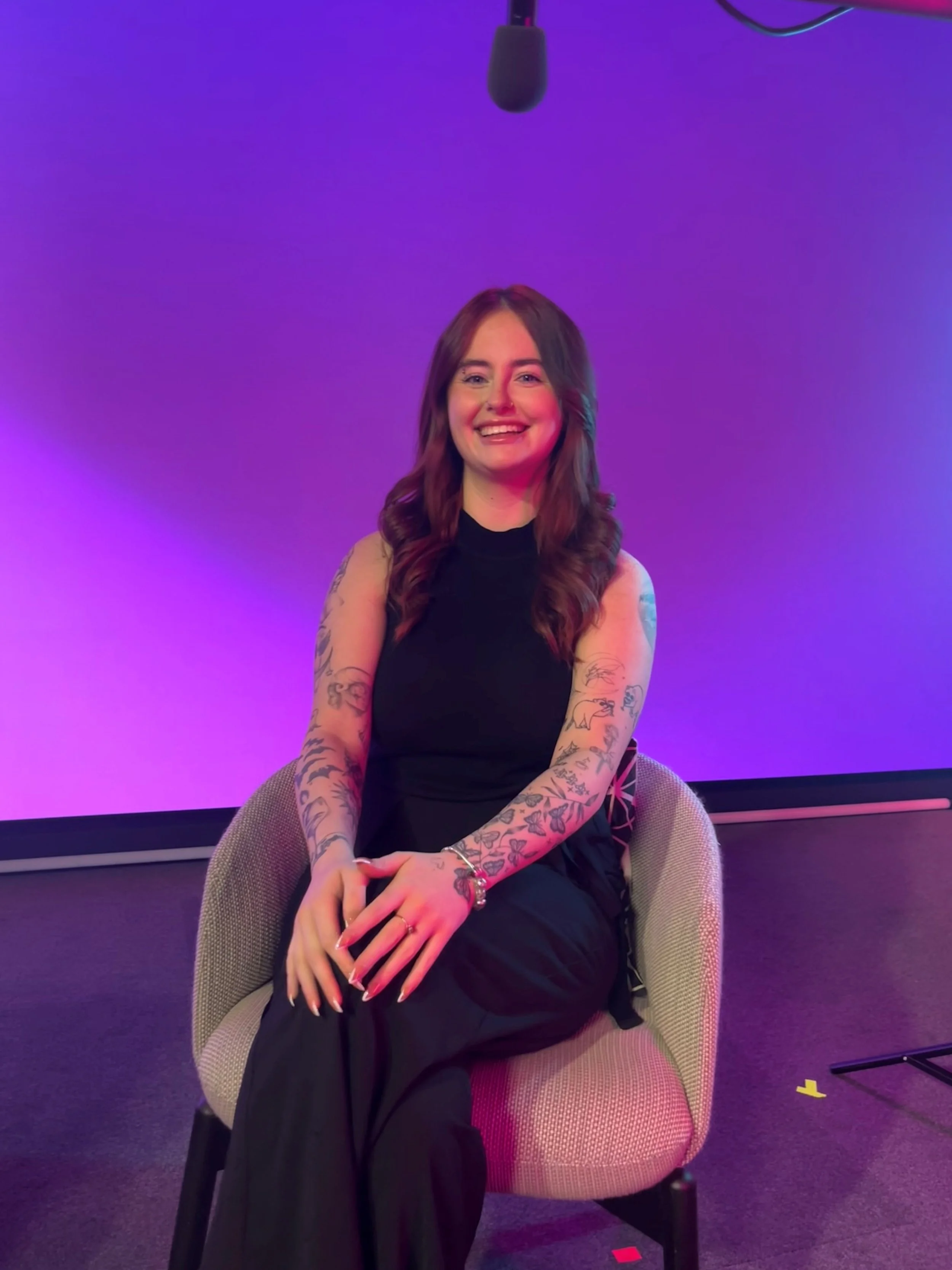 Photo of Jessie Wright who has long dark wavy hair smiling wearing a black dress, sitting on a chair on a stage with purple background.