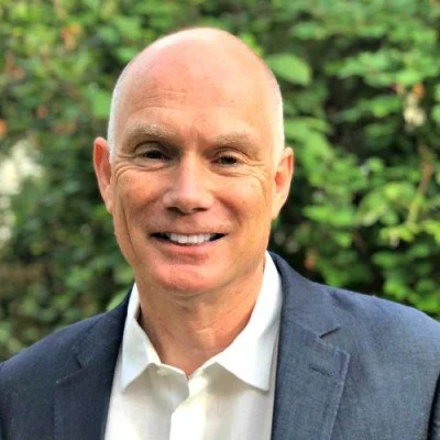 Headshot of Iain Wilkie smiling with a bald head, wearing a white shirt and dark grey suit jacket