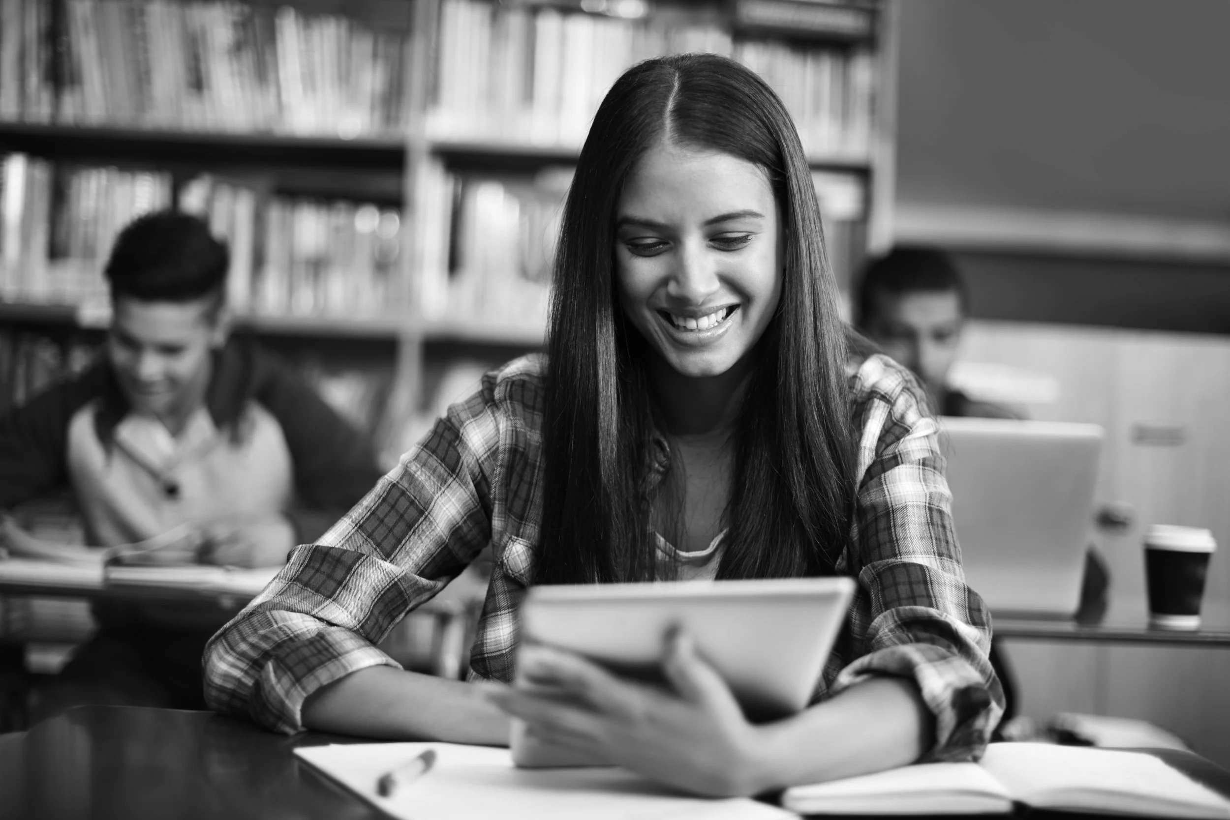 Young woman with long dark hair and checked shirt sitting in a library smiling looking down at a tablet.