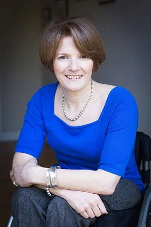 Photo of Helen Cooke sitting in her wheelchair leaning forward wearing a bright blue top, hair in a short bob and smiling.