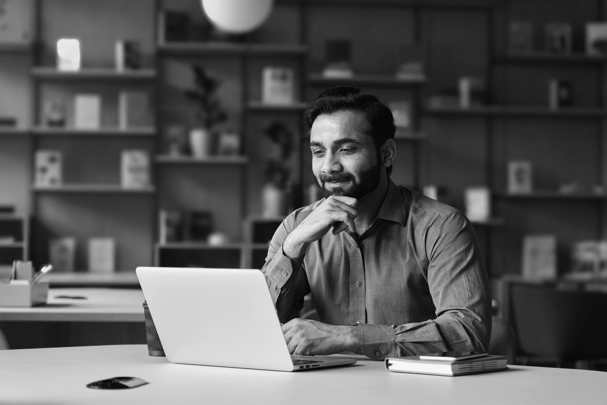 Man with dark hair and beard sitting looking at a laptop with his hand under his chin