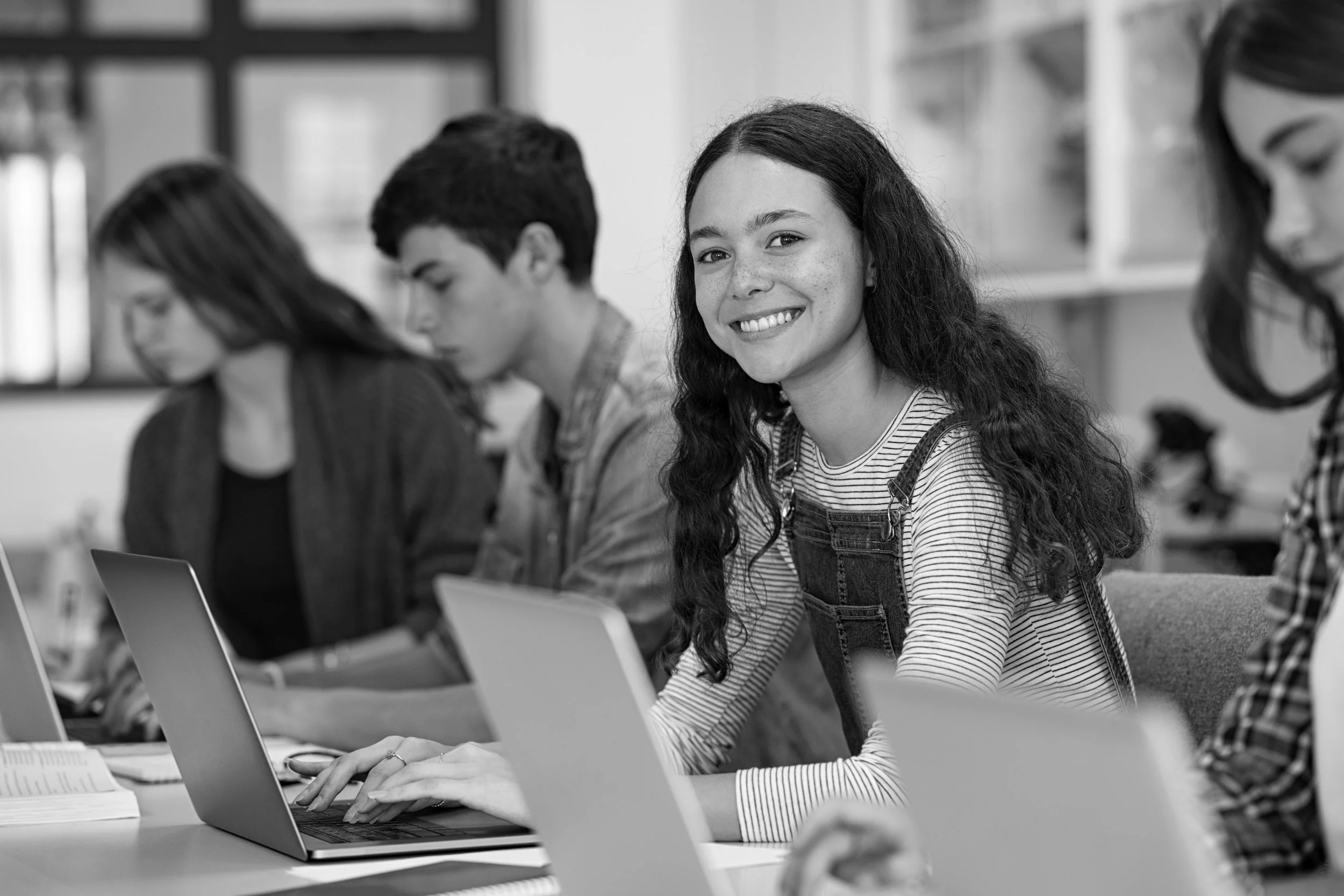 Young woman with long wavy hair smiling sitting at a desk  with her laptop and other students  on either side