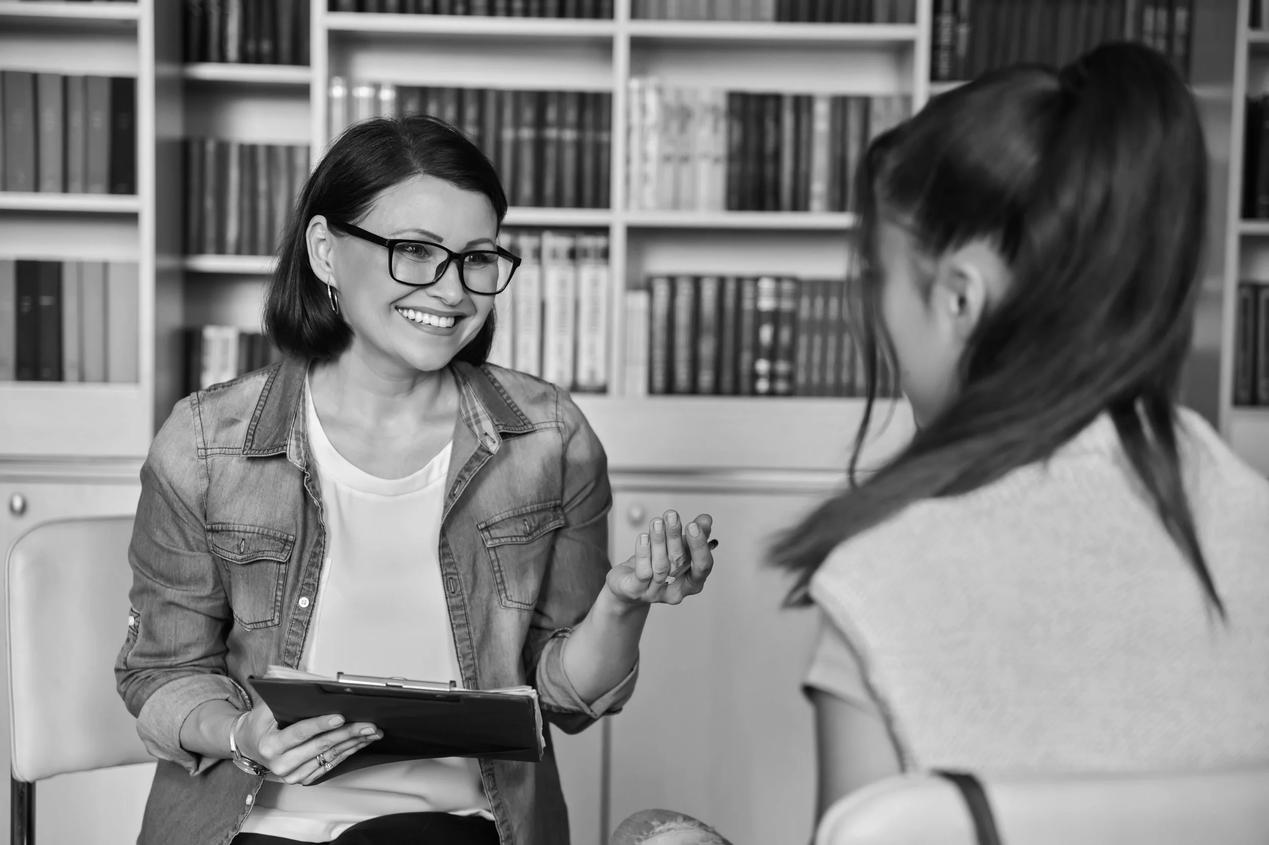 Woman with bobbed dark hair and glasses sitting taking to a young woman with dark hair in a pony tail with her back to the camera.