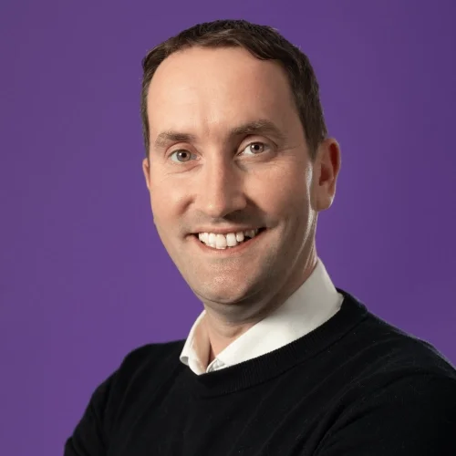 Headshot of Martin Kavanagh with  short dark hair smiling wearing a white shirt and dark jumper with a purple background