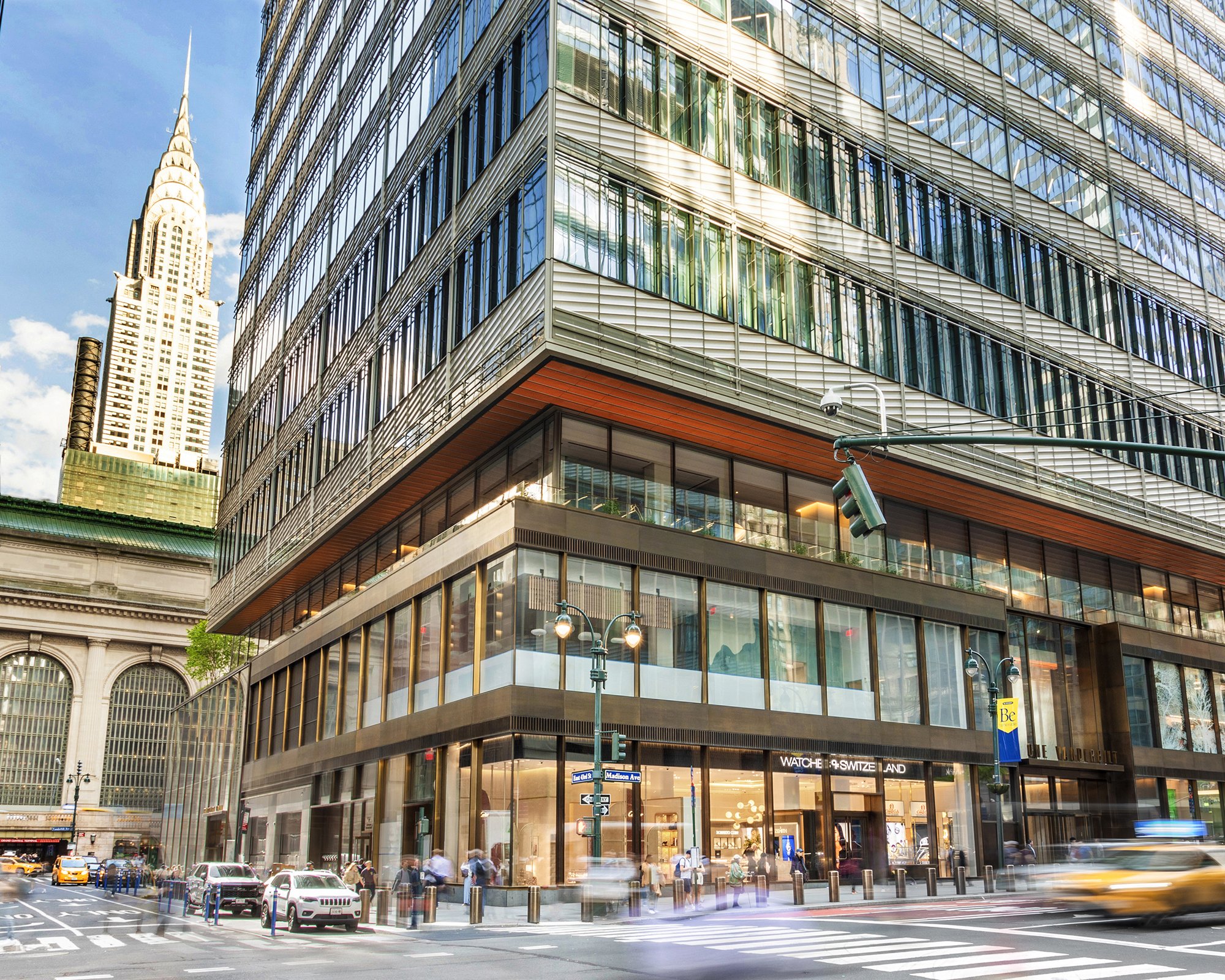 Modern glass office building at a busy city corner with Madison Avenue signs, featuring reflections and a view of the Chrysler Building in the background, with pedestrians and cars on the street.