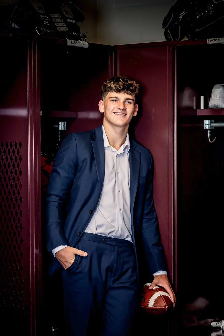 A young man in a suit smiling, standing inside a maroon locker room, holding a football in his right hand.