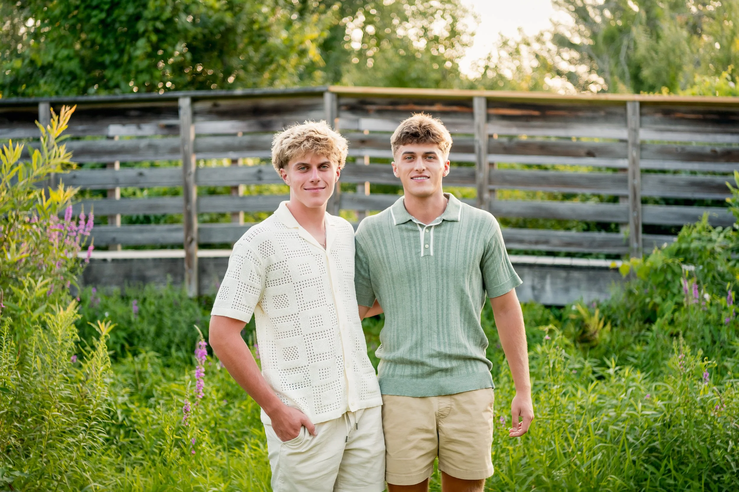 Two young men standing outdoors in a garden with green foliage and purple flowers, smiling at the camera during the daytime.
