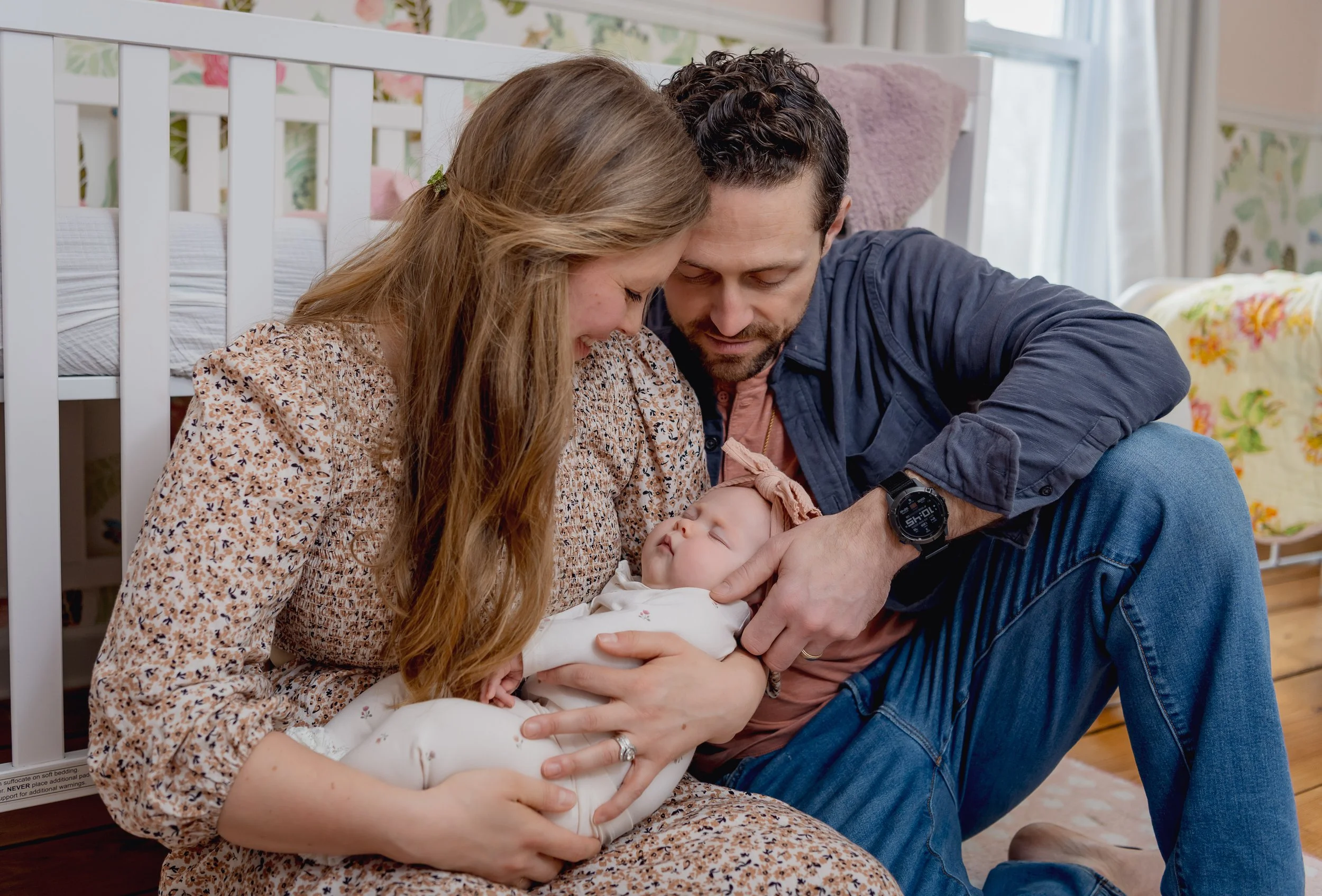 Parents with a newborn baby in a cozy nursery, sitting on the floor under a crib, smiling and looking at their baby.