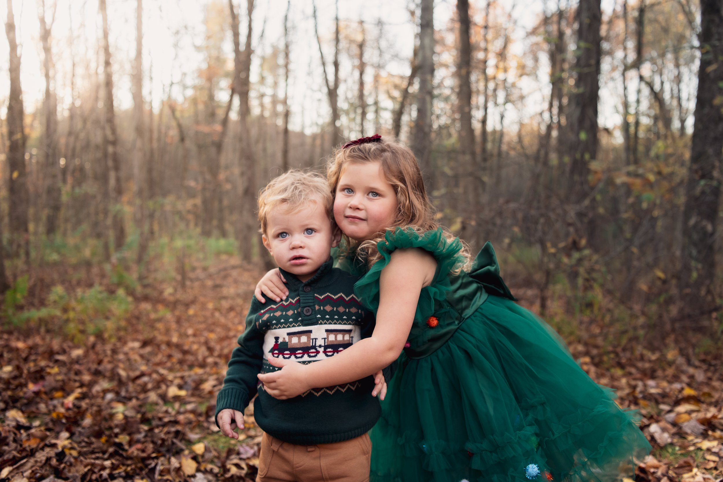 Two children, a boy and a girl, standing together in a wooded area with fallen autumn leaves. The girl is wearing a green dress with tulle, and the boy is wearing a festive sweater with a train pattern. They are close to each other, with the girl hug
